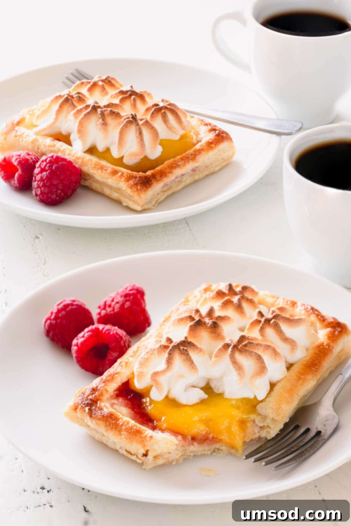 Raspberry lemon meringue tartlets on white plates with fresh raspberries and cups of black coffee.