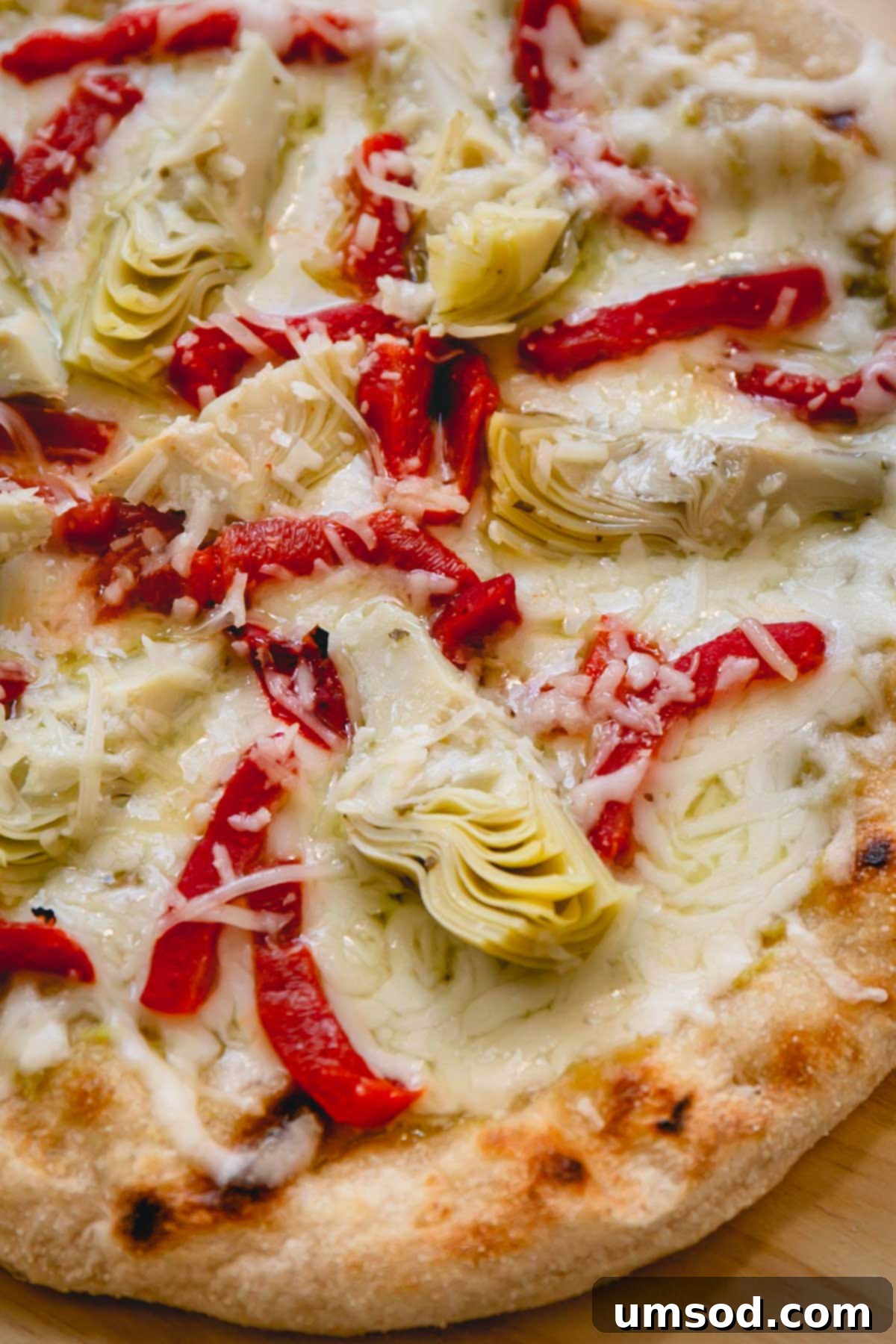 A close-up shot of a whole grilled artichoke pizza, showcasing its melted mozzarella, roasted red peppers, and vibrant pesto swirls.