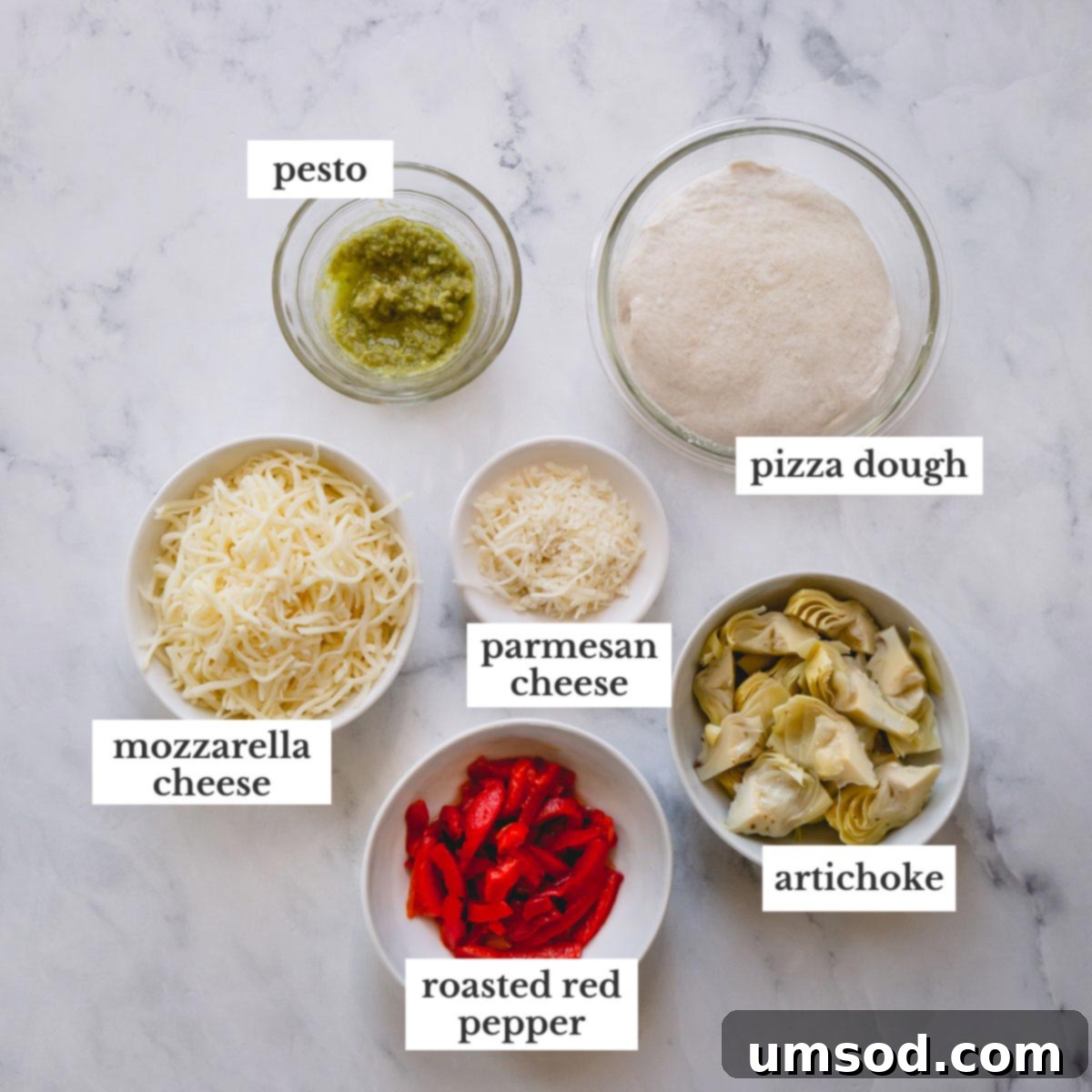 A flat lay of the fresh ingredients for grilled artichoke pizza, including pizza dough, a jar of artichoke hearts, roasted red peppers, mozzarella, pesto, and parmesan cheese.