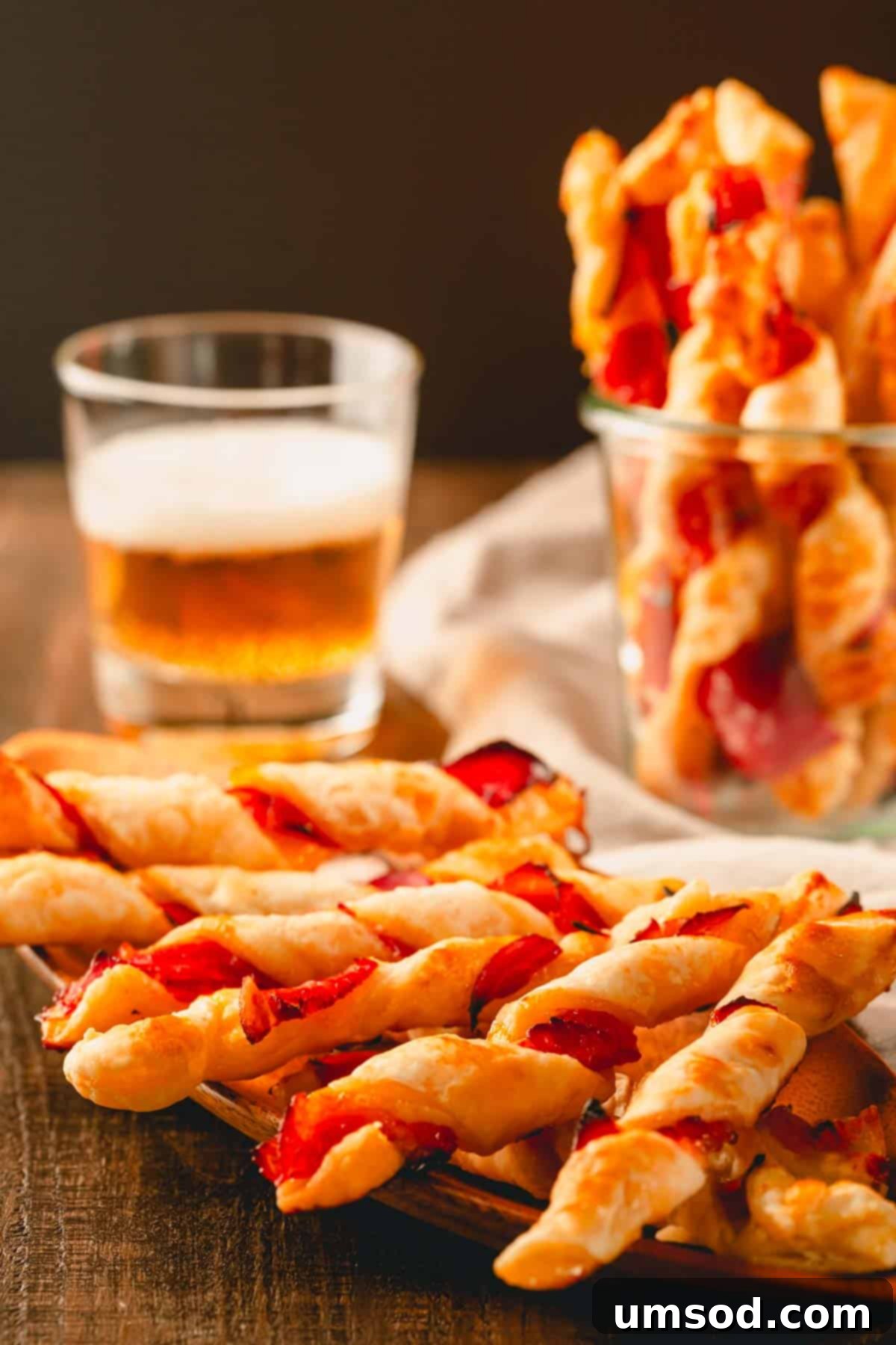 Prosciutto puff pastry twists on a plate and in a glass cup with a glass of beer in the background, suggesting a perfect pairing.