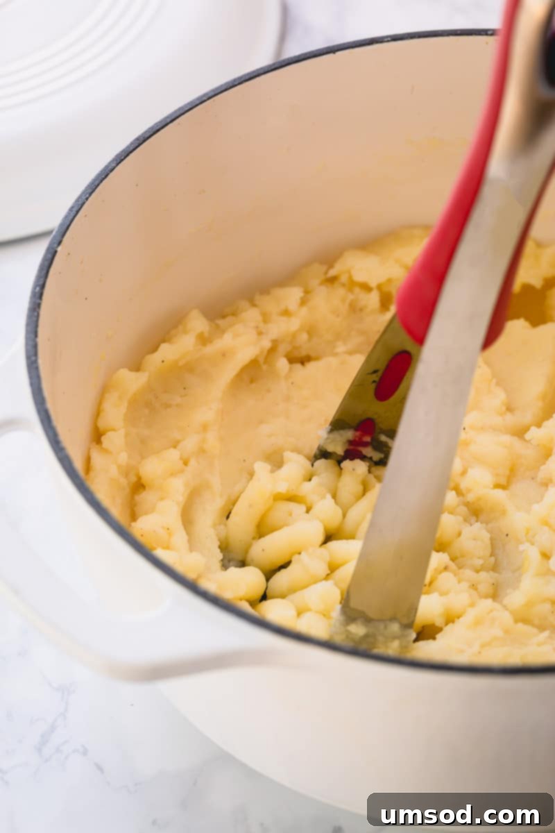 Ultimate Loaded Potato Bake 5 Mashed potatoes in a white Dutch oven with a potato masher in it.