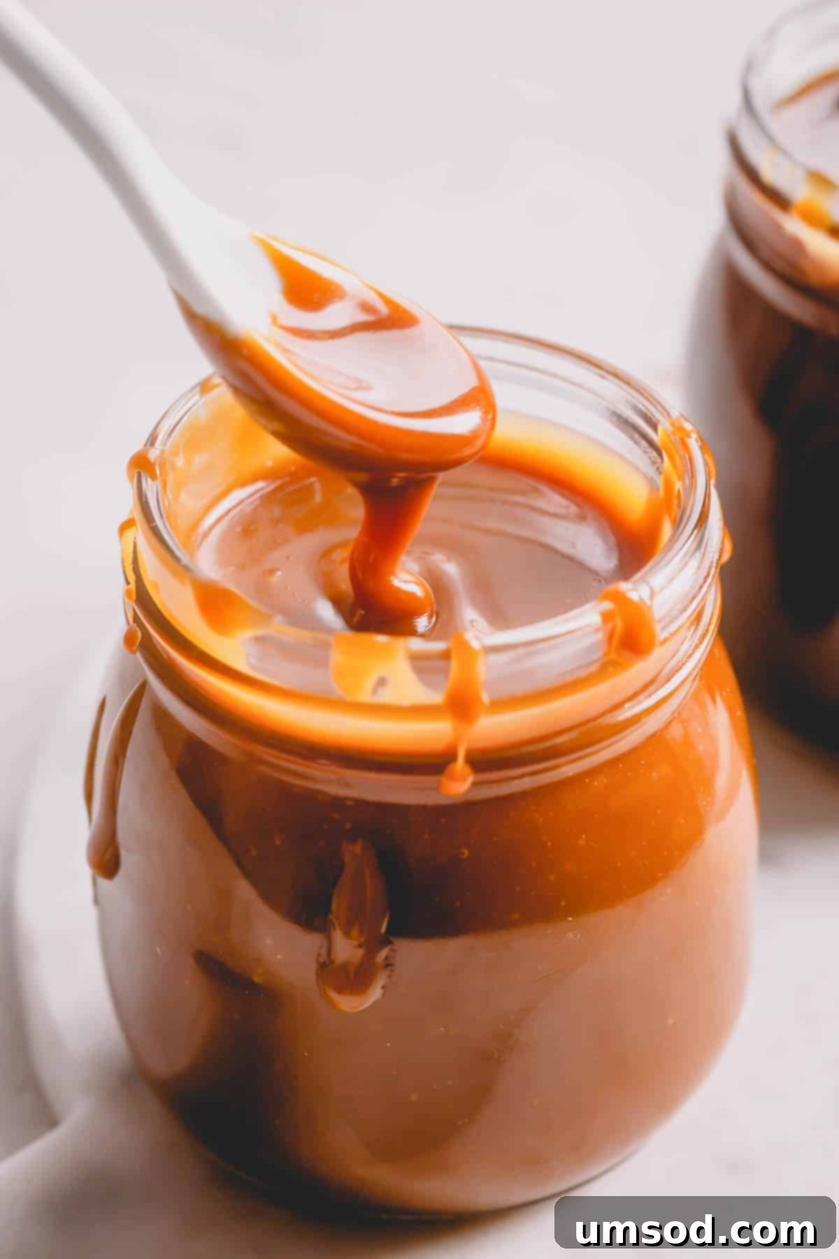 Homemade Caramel Sauce Dry Method Guide 2 A jar of golden homemade caramel sauce with a spoon lifting a scoop out, showcasing its rich, thick texture.