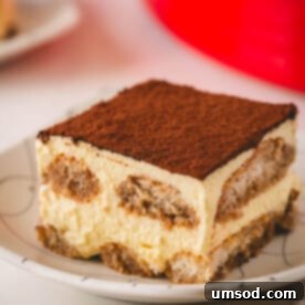 A perfectly portioned square of traditional tiramisu on a white plate, revealing its elegant layers of coffee-soaked ladyfingers and creamy mascarpone.