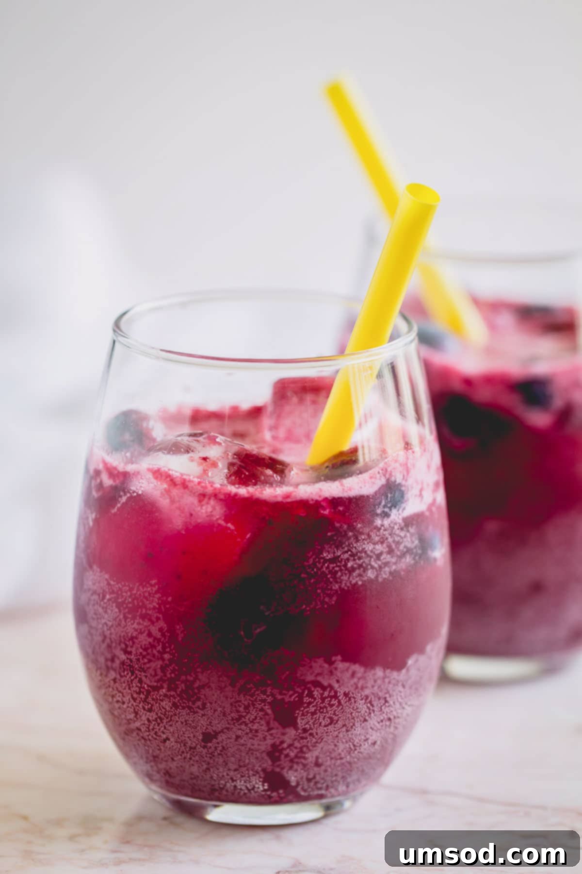 A tall glass of sparkling blueberry lemonade with a yellow straw, garnished with fresh blueberries and a lemon wedge.