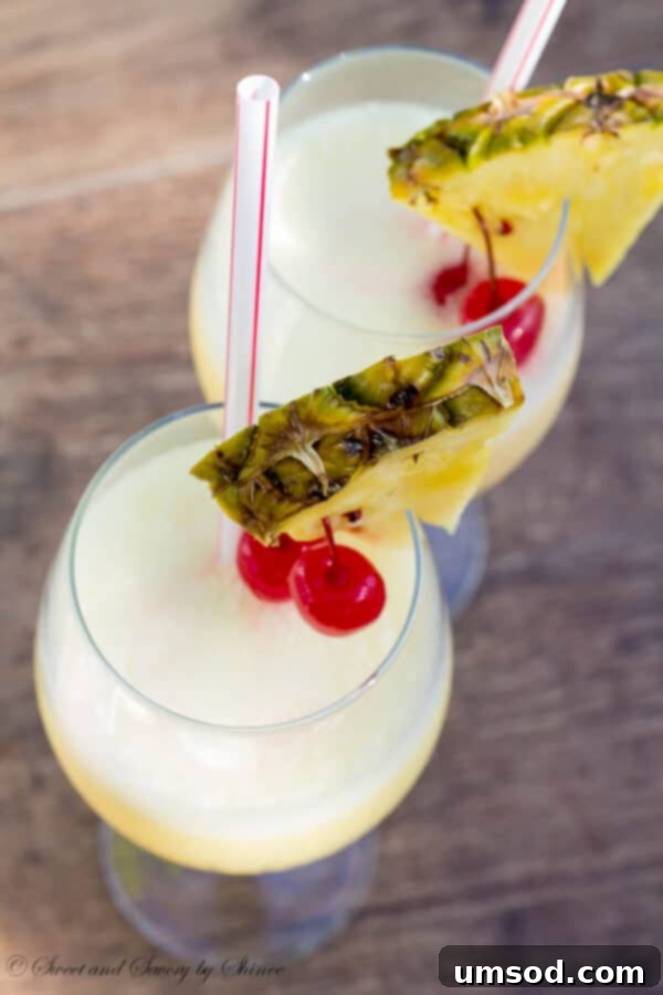 Two glasses of Pina Colada garnished with fresh pineapple and cherries, set against a blurred tropical background