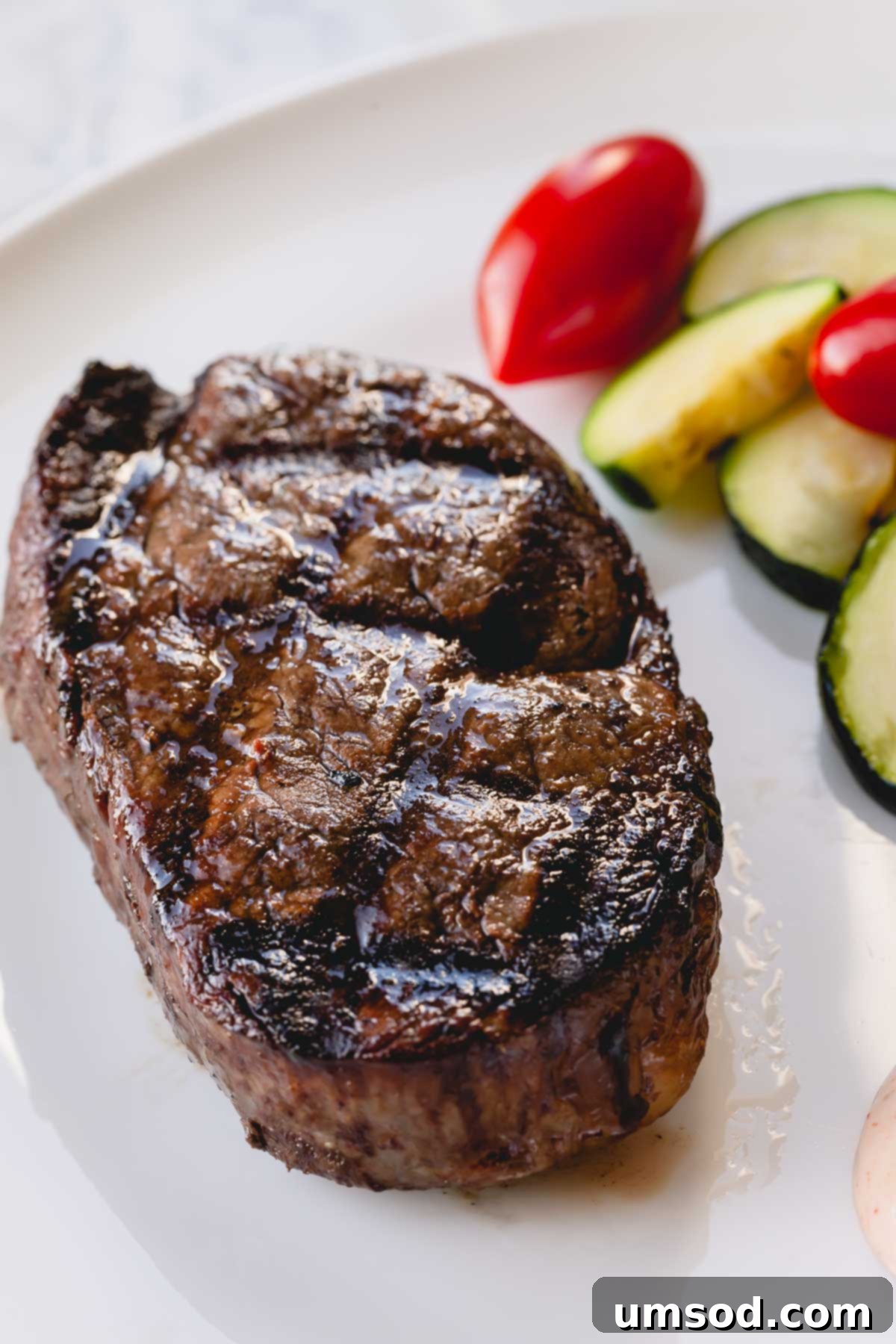 Effortless Grilled Filet Mignon 3 A whole grilled filet mignon on a white plate, beautifully garnished with vibrant zucchini and cherry tomatoes on the side.