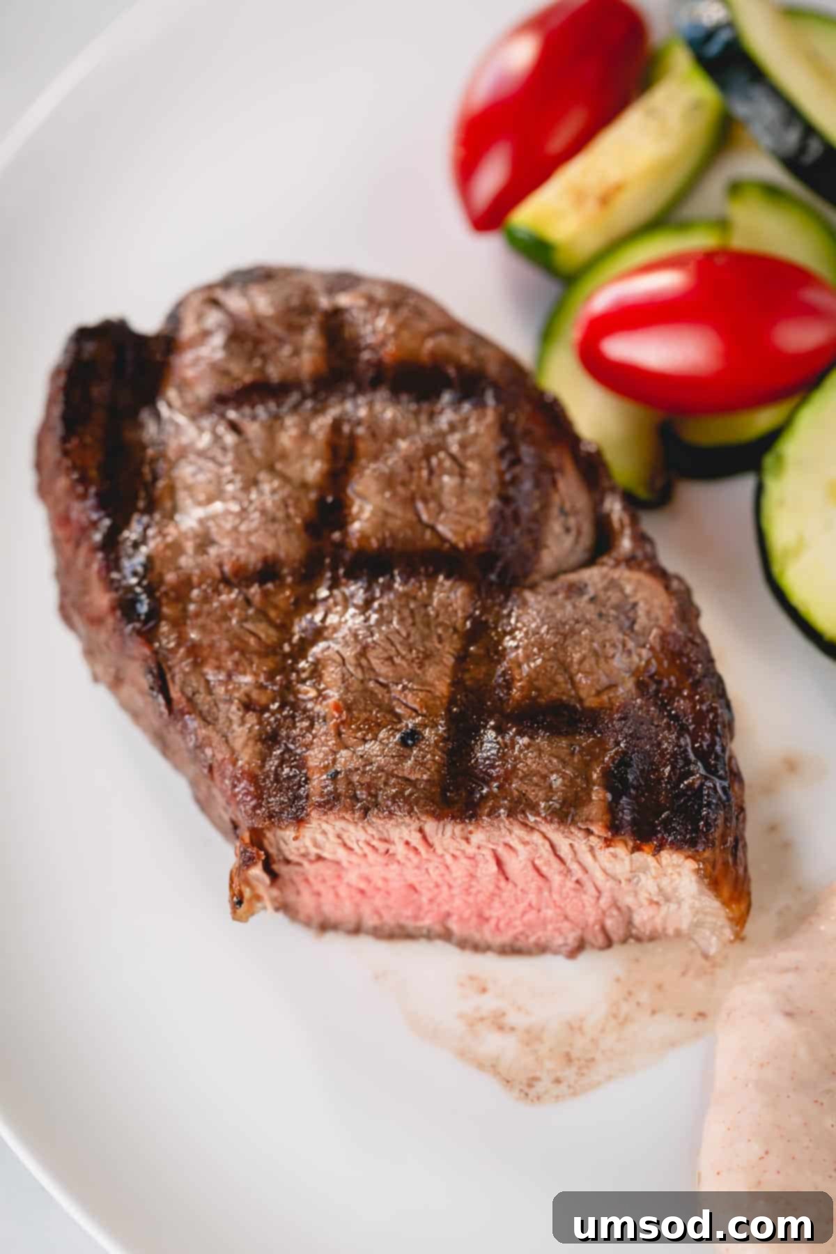 Effortless Grilled Filet Mignon 4 A whole grilled filet mignon on a white plate with zucchini and cherry tomatoes on the side, highlighting the delicious meal.
