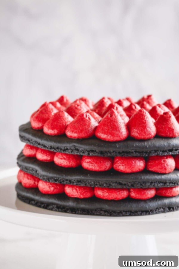 A 3-layer black macaron cake being assembled with red cream cheese filling on a white cake stand.