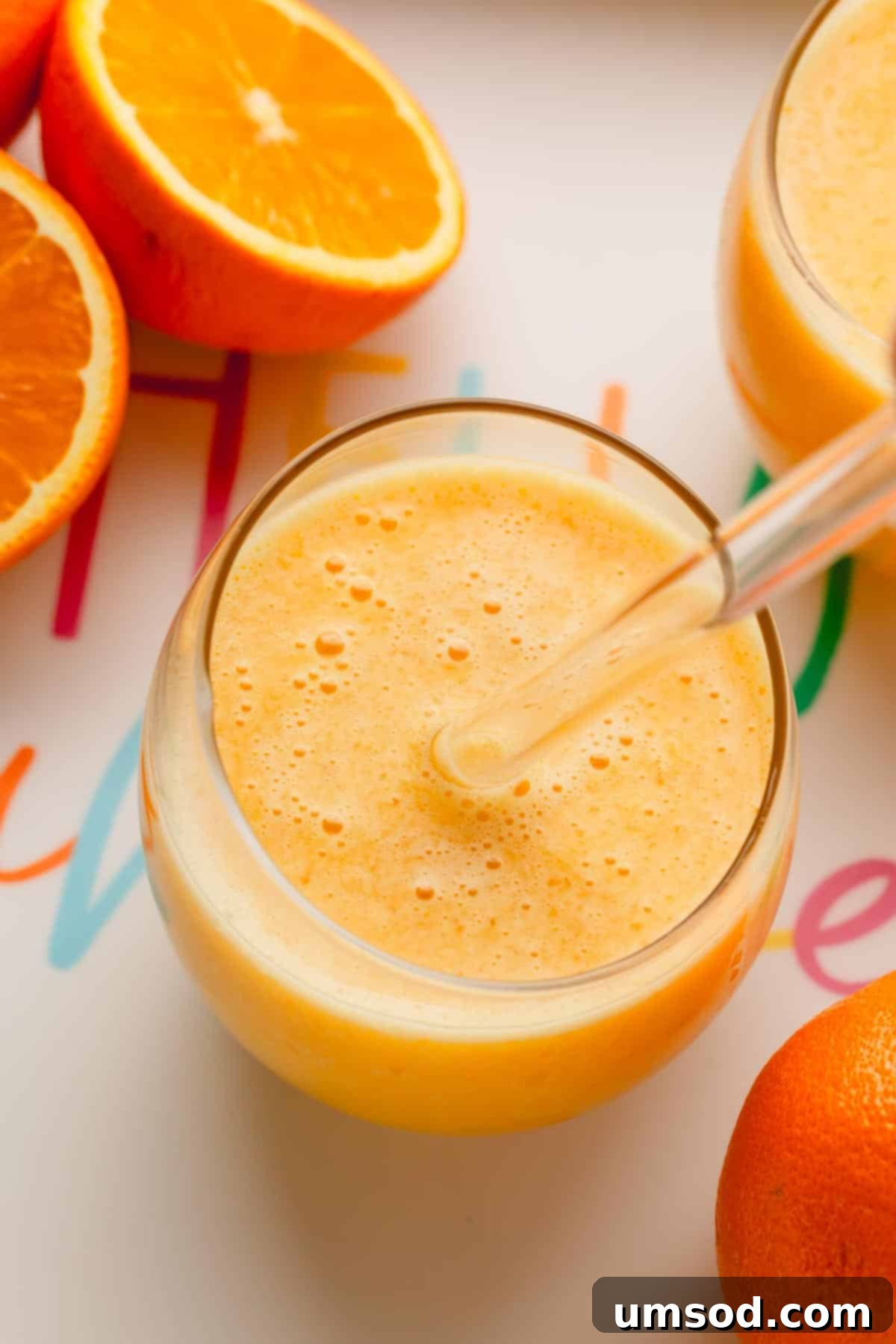 A glass of coconut orange Julius with a glass straw sticking out and oranges around it.