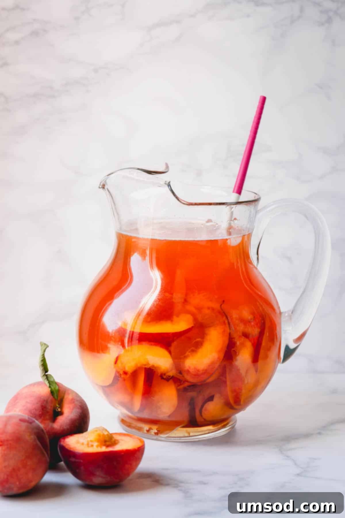 Peachy Keen Iced Tea 5 A large glass pitcher filled to the brim with homemade peach iced tea, garnished with fresh peach slices and mint, against a light background.