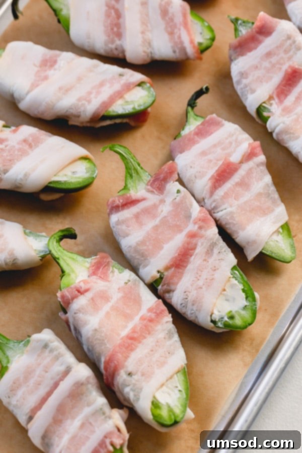 Bacon wrapped jalapeno poppers arranged on a parchment-lined baking sheet, ready to go into the oven.