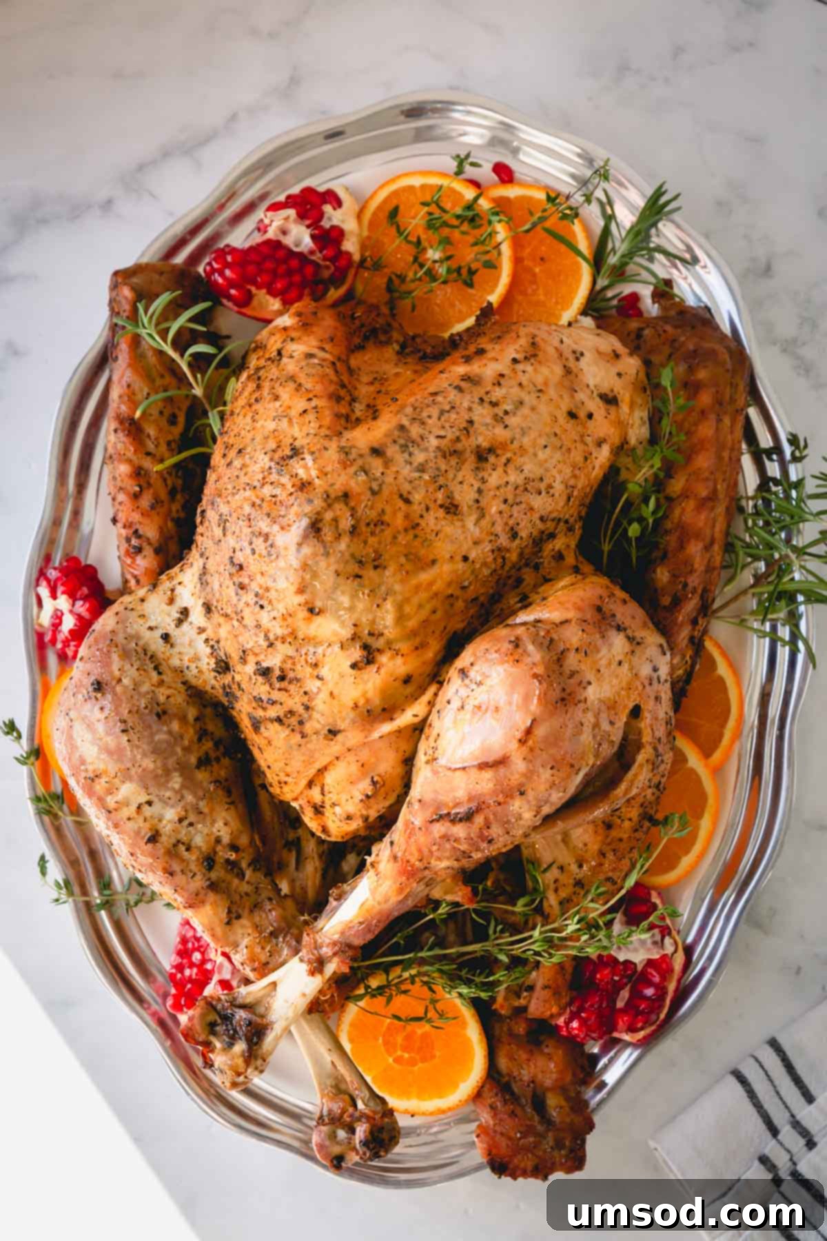 Whole roast turkey on a silver platter garnished with vibrant orange slices, pomegranate halves, and fresh herbs.