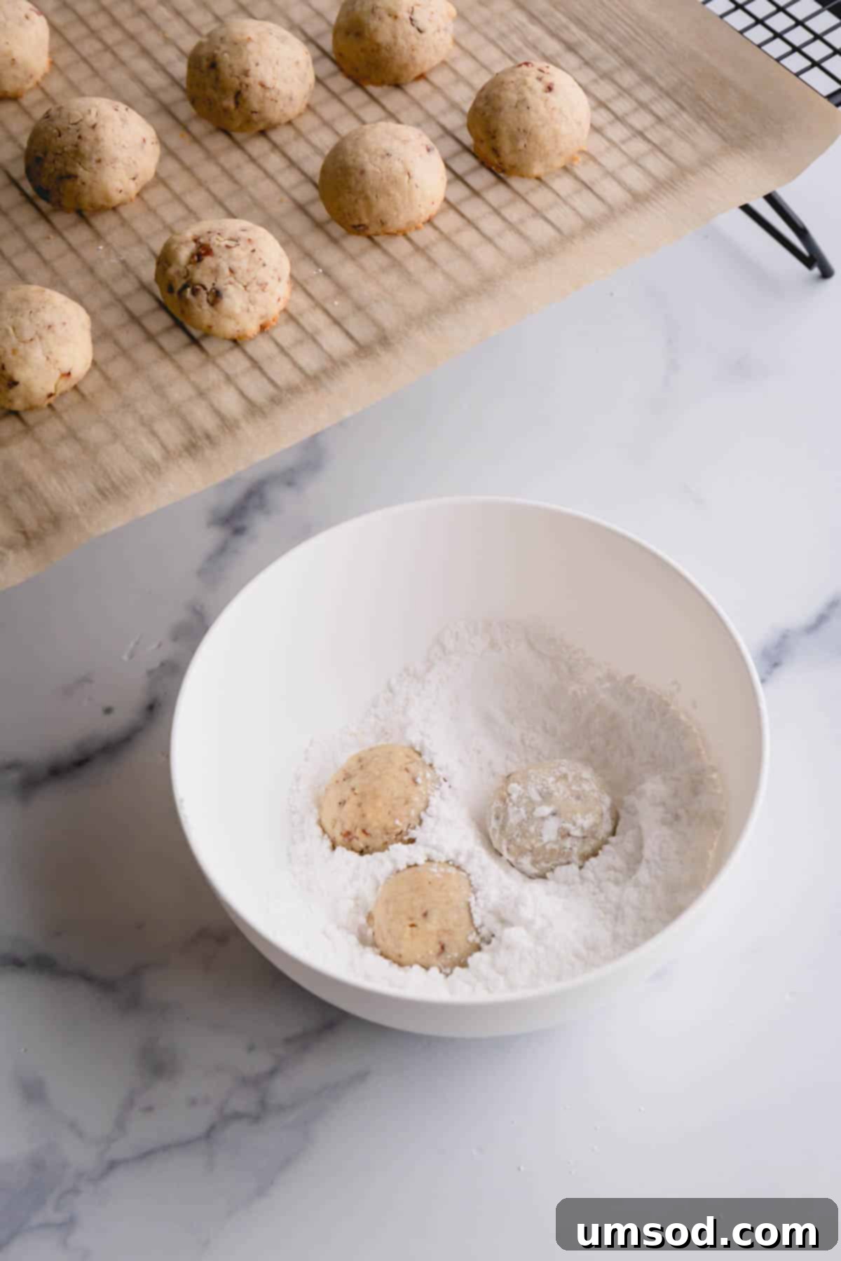 A warm snowball cookie being gently rolled in a bowl of powdered sugar for its first coating.