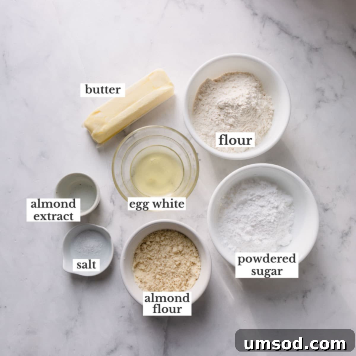 A collection of ingredients laid out on a surface, including butter, sugar, flour, and extracts, ready for making almond spritz cookies.