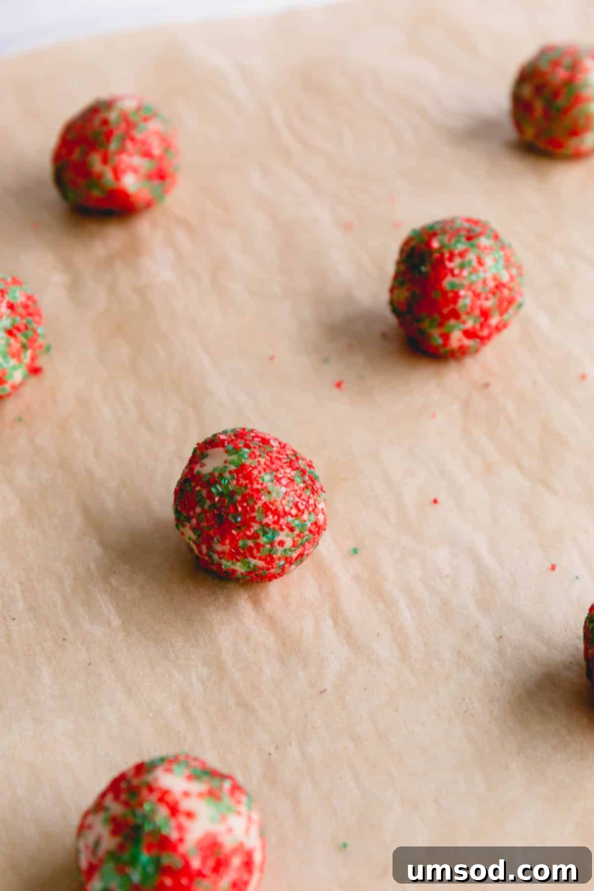 Tek Hamurdan 6dan Fazla Lezzet: Efsanevi Tereyağlı Kurabiye Hamuru 18 Small cookie dough balls being rolled in festive red and green colored sugars on a parchment-lined surface.