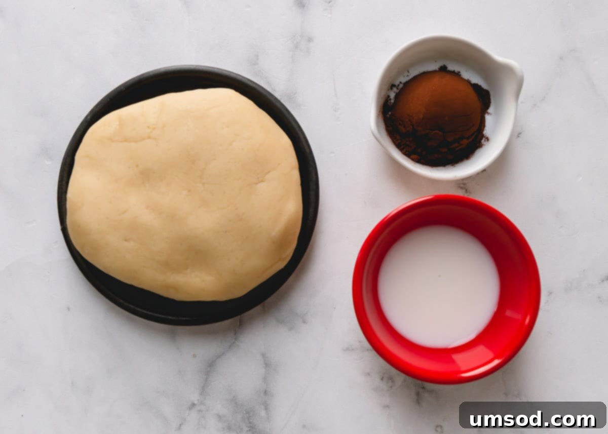 Tek Hamurdan 6dan Fazla Lezzet: Efsanevi Tereyağlı Kurabiye Hamuru 6 A cookie dough ball, a small bowl of cocoa powder, and a measuring spoon of milk, prepared for making chocolate cutout cookies.