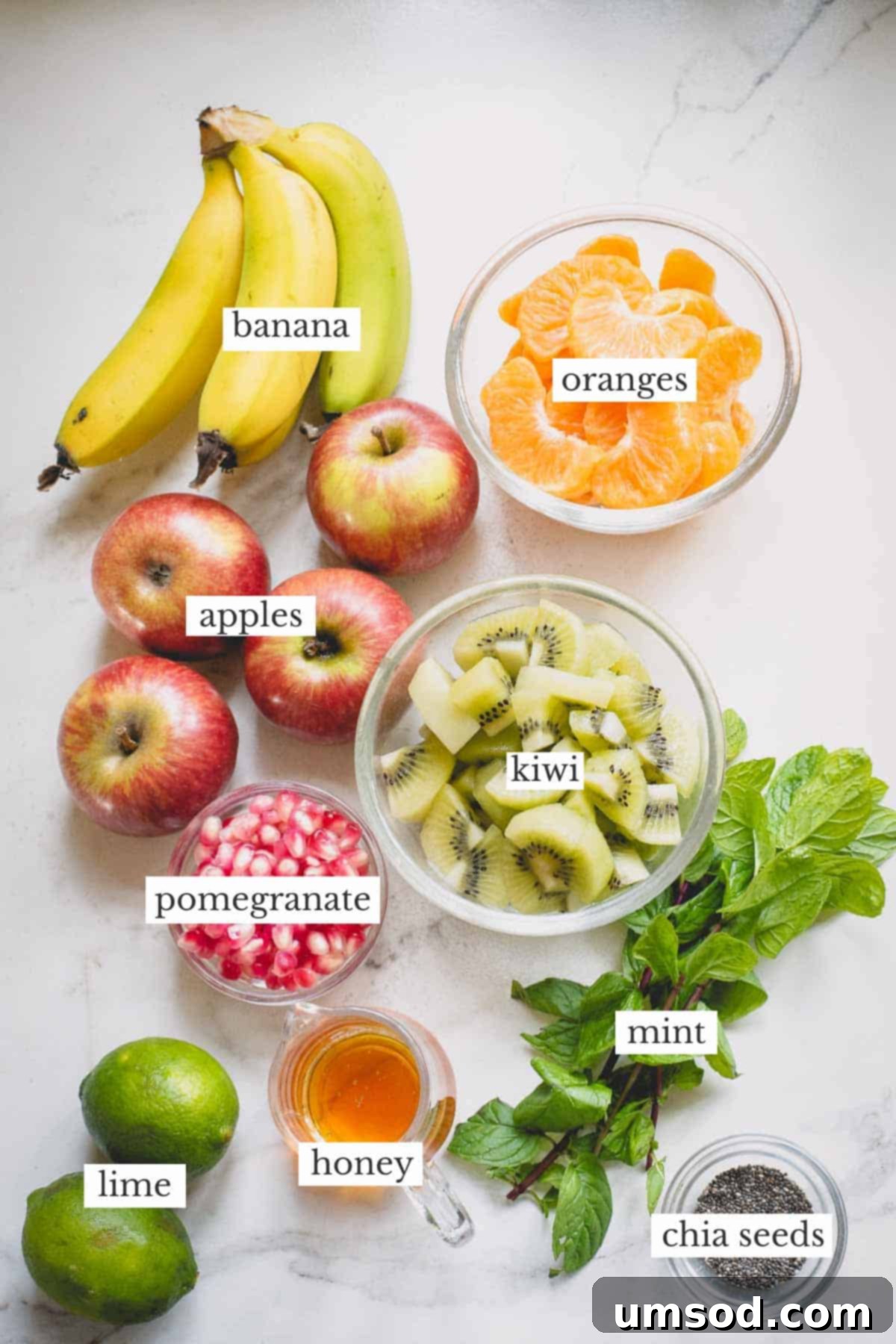 Fresh Fruit Medley 3 A selection of fresh ingredients including bananas, apples, oranges, kiwi, mint leaves, limes, and a jar of honey, laid out on a surface.