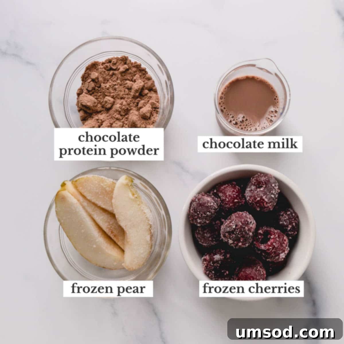 A curated selection of key ingredients for the Black Forest Smoothie Bowl: chocolate protein powder, a bottle of chocolate milk, a fresh pear, and a bowl of frozen cherries, arranged for cooking.