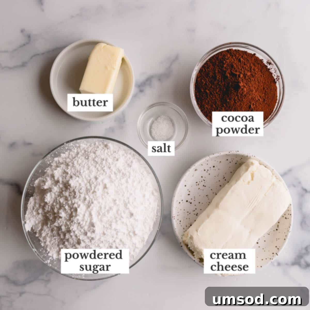 Key ingredients for chocolate cream cheese frosting: a block of cream cheese, a stick of butter, a bowl of cocoa powder, and a bowl of powdered sugar.