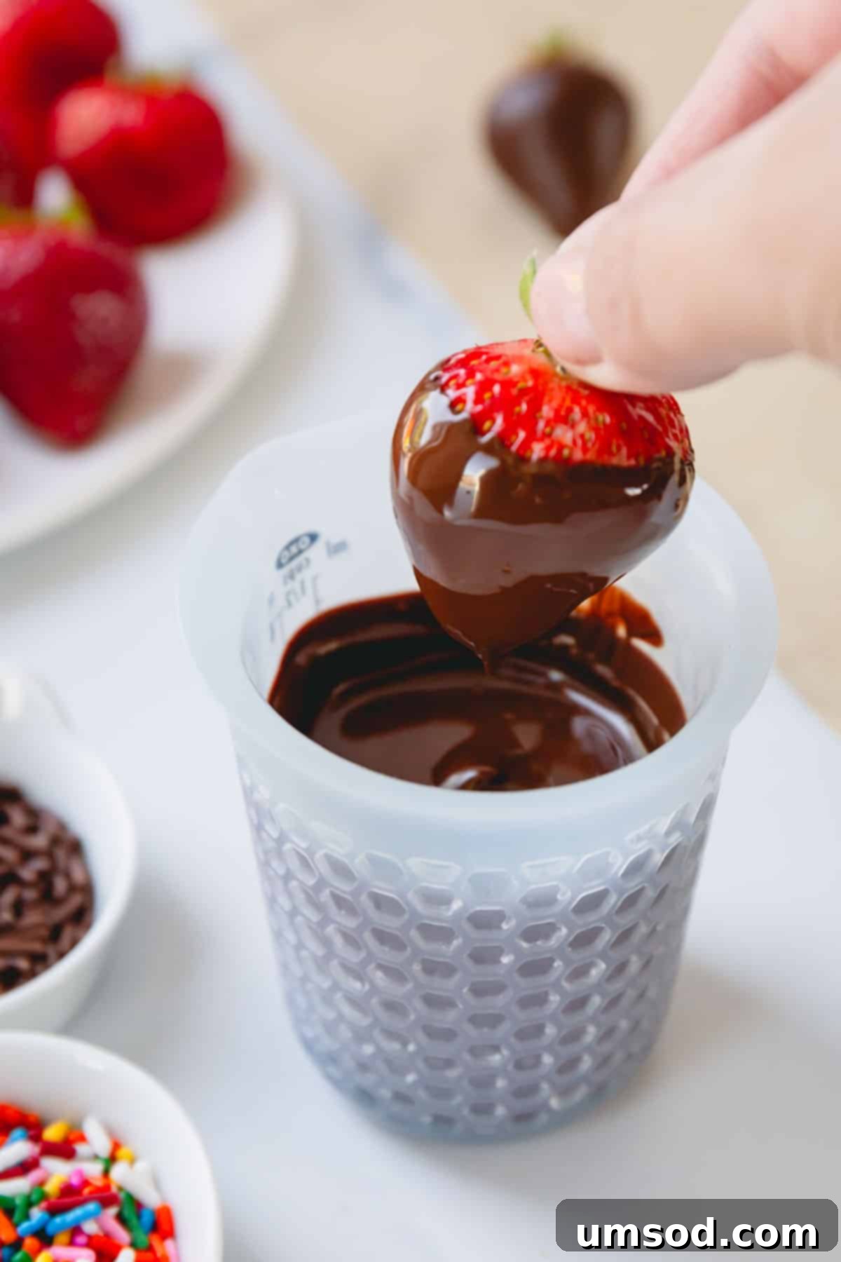 A hand holding a chocolate covered strawberry by the stem over a cup of melted chocolate.