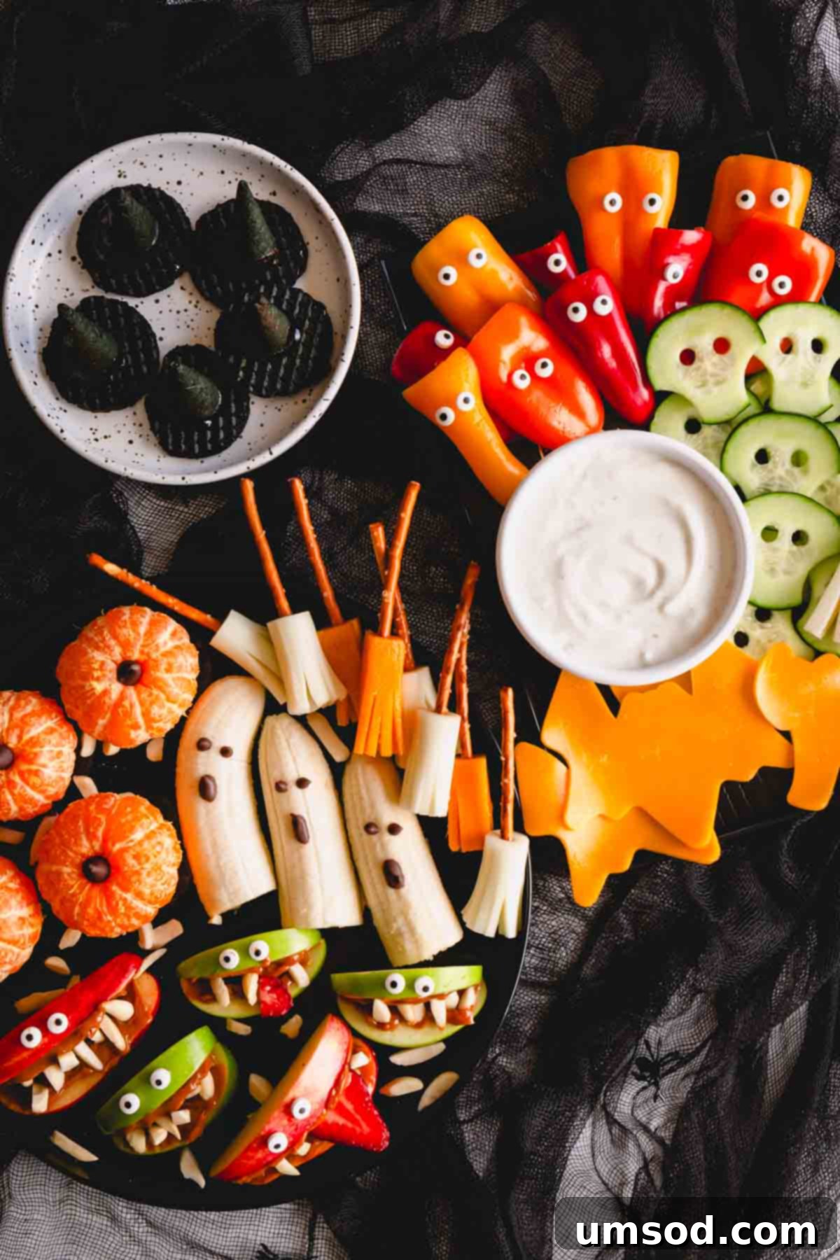A vibrant and healthy Halloween snack board featuring ghost-nanas, pumpkin oranges, and apple monster mouths arranged creatively for kids.