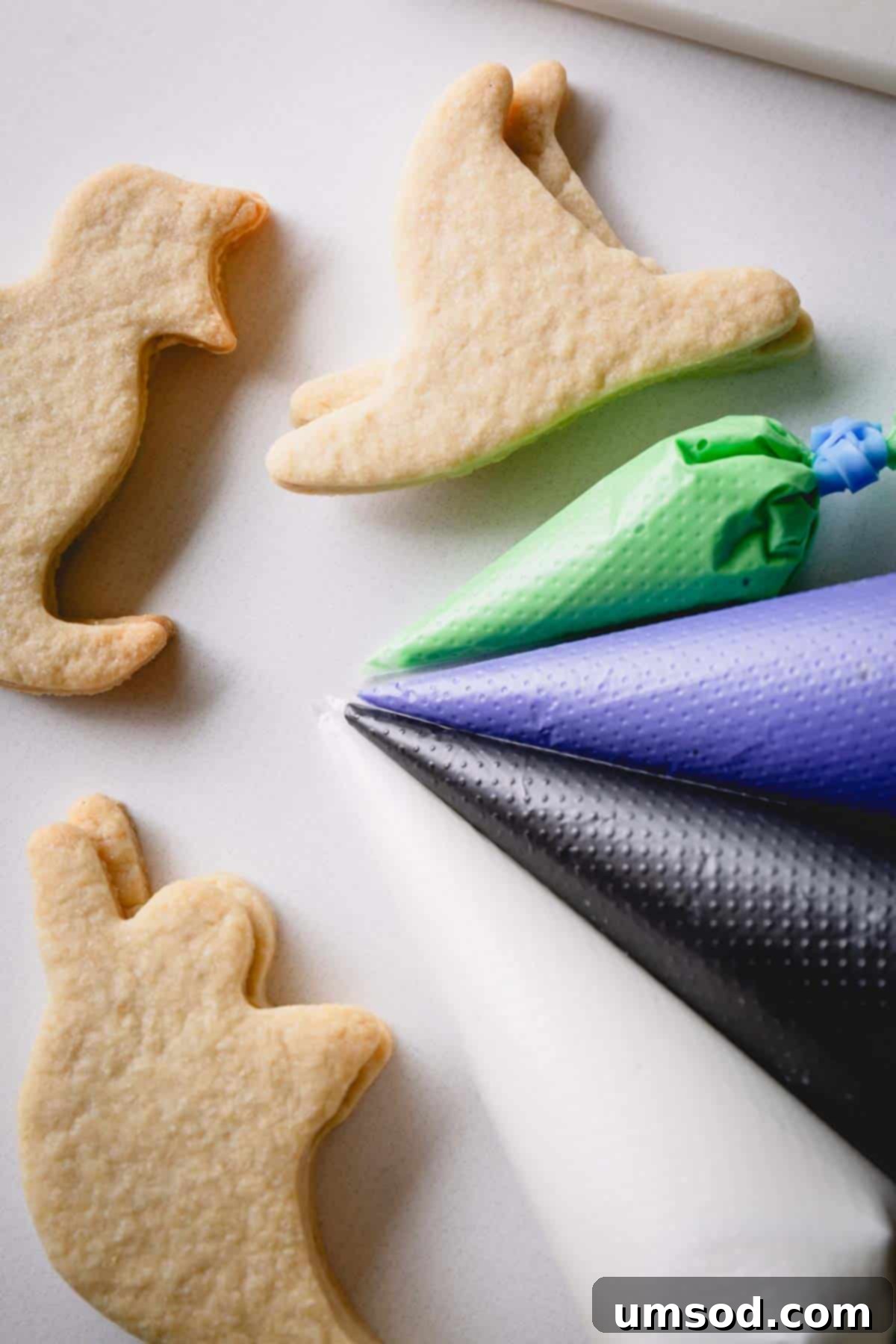 Baked Halloween sugar cookies next to piping bags full of green, purple, black, and white icing.