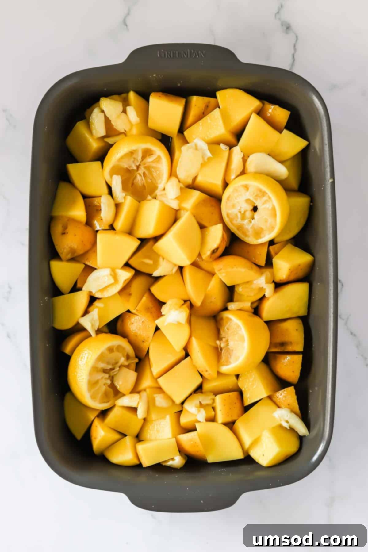 A baking dish filled with chopped potatoes, smashed garlic cloves, and halved lemons, ready for seasoning.