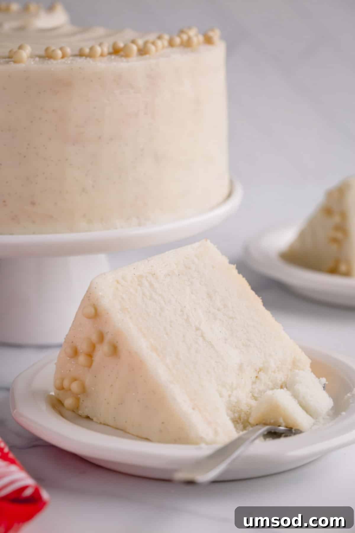Fluffy White Cake Perfection 14 A delectable slice of classic white cake on a plate with a fork, while the beautifully frosted whole cake stands in the background.