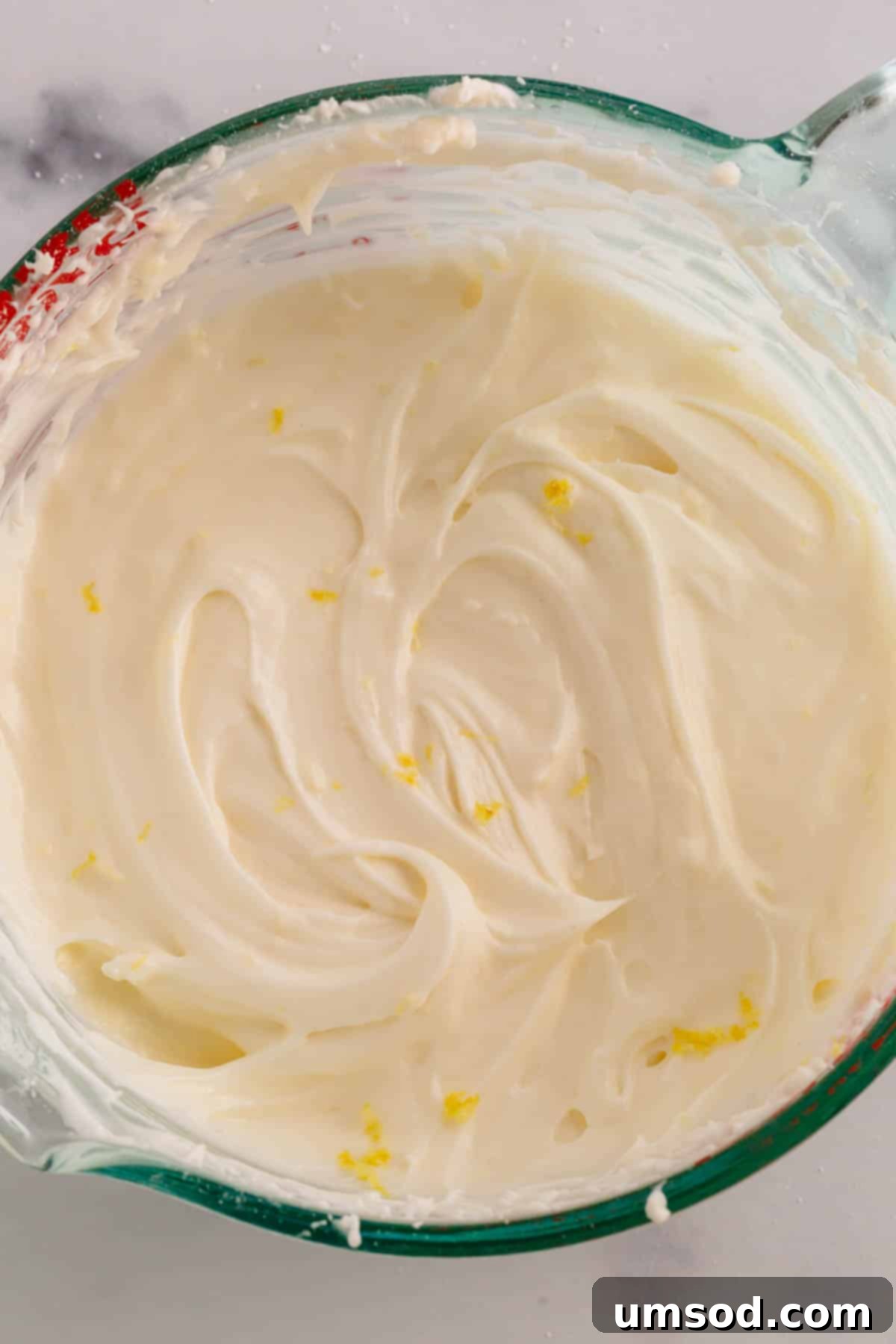 A liquid measuring cup full of lemon cream cheese frosting with flakes of lemon zest.