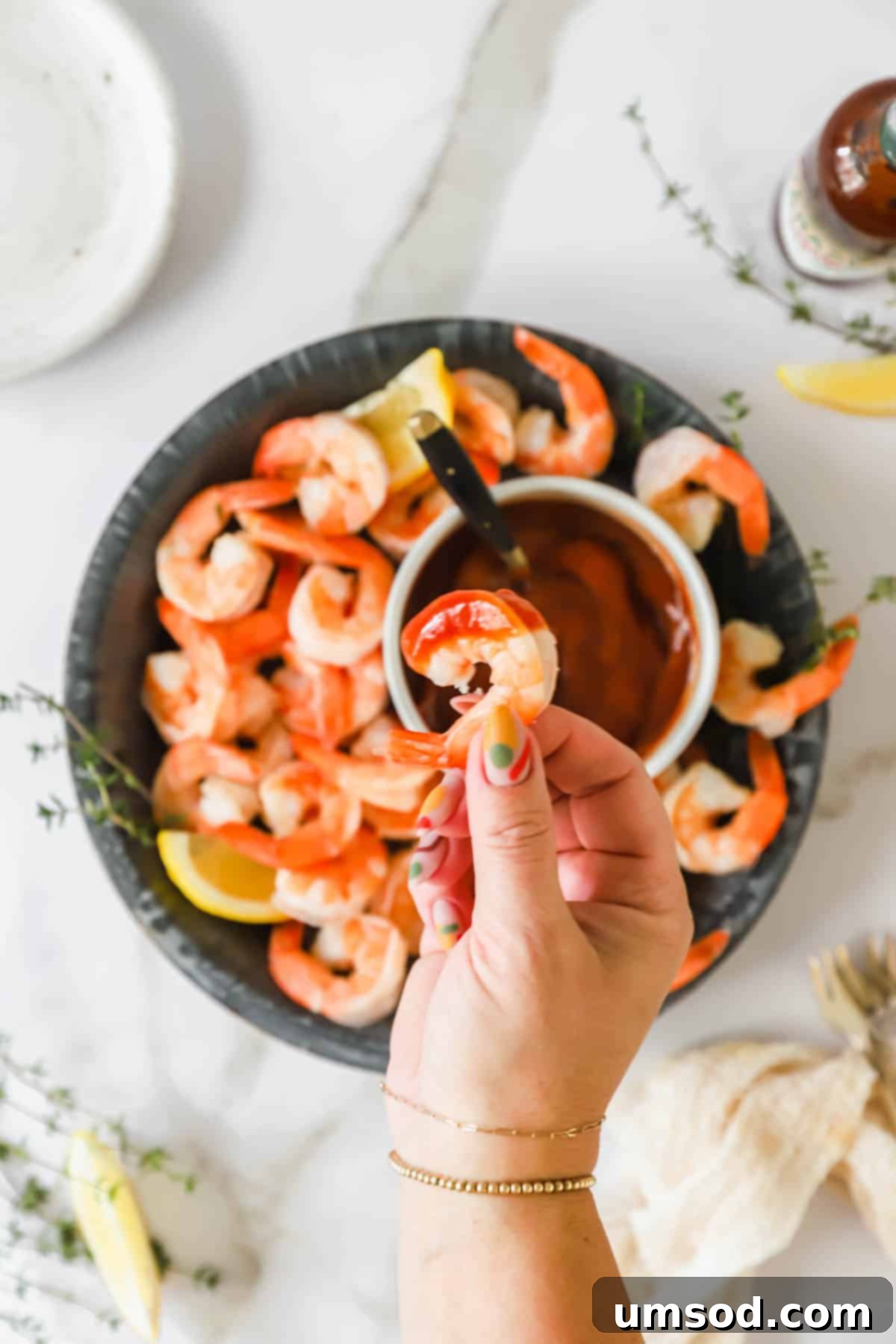 A hand gracefully holds a cocktail shrimp, poised just above a swirl of rich, red cocktail sauce, ready for dipping.