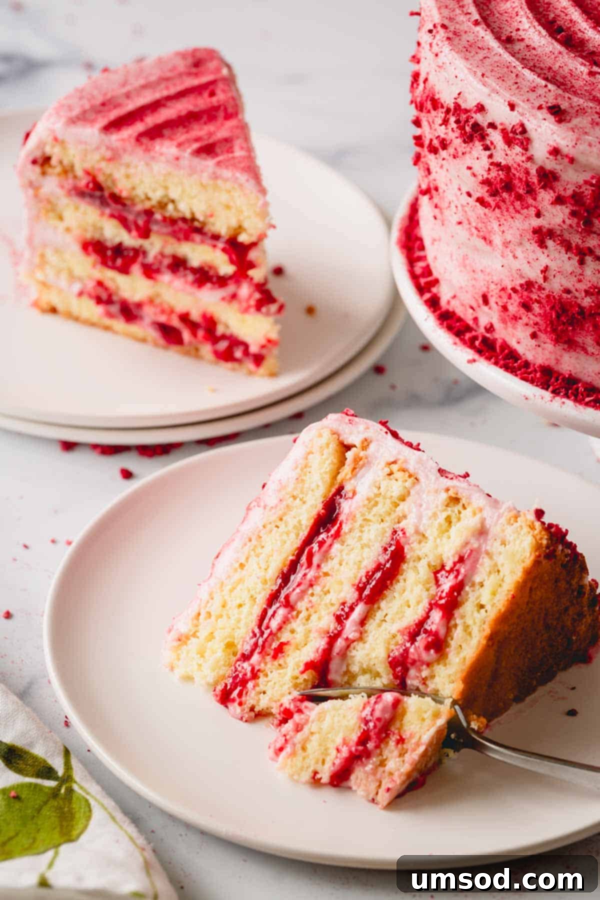 Luscious Raspberry Layer Cake 12 Two generous slices of raspberry layer cake presented on plates, positioned beside the whole cake, with a fork poised to take a bite from one of the slices.