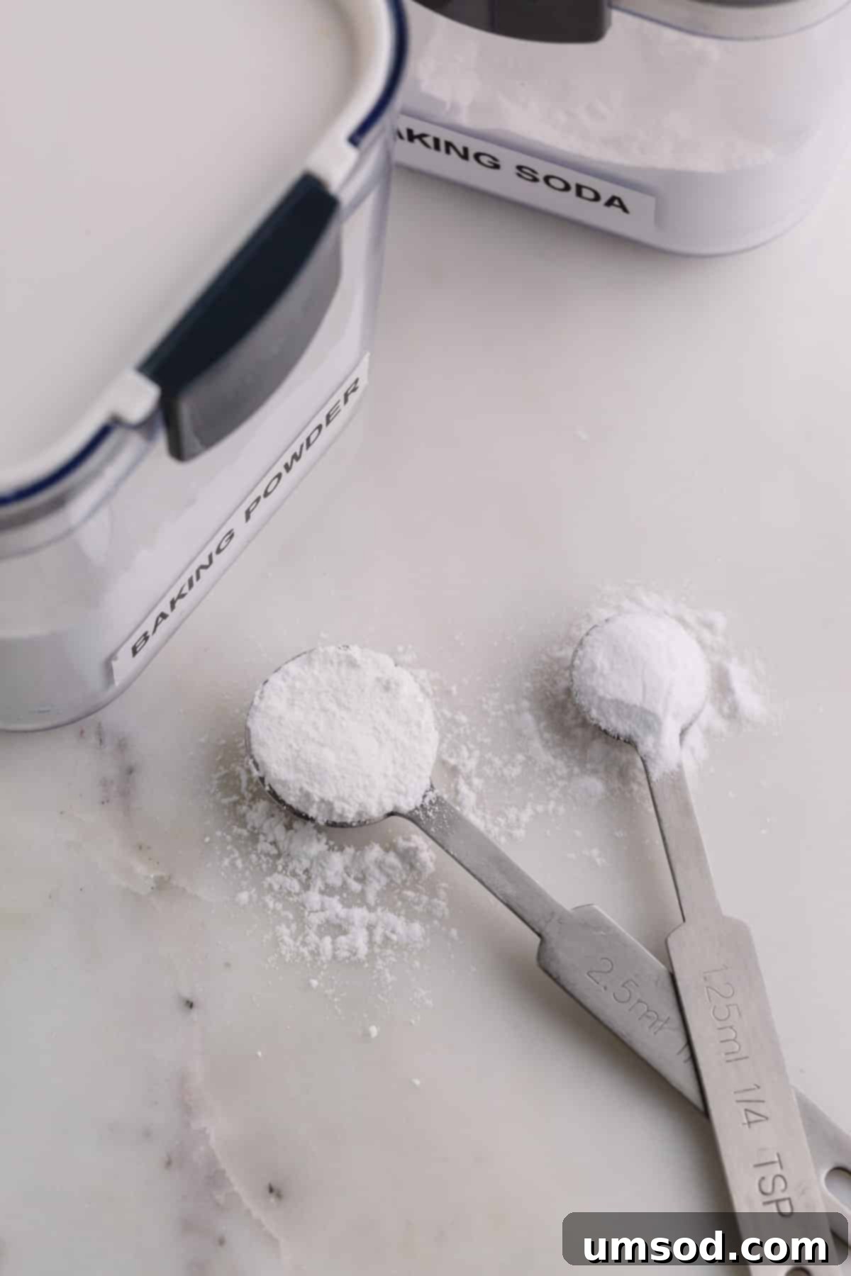 Two measuring spoons on a marble background, one with baking powder and one with baking soda, illustrating their distinct appearance.