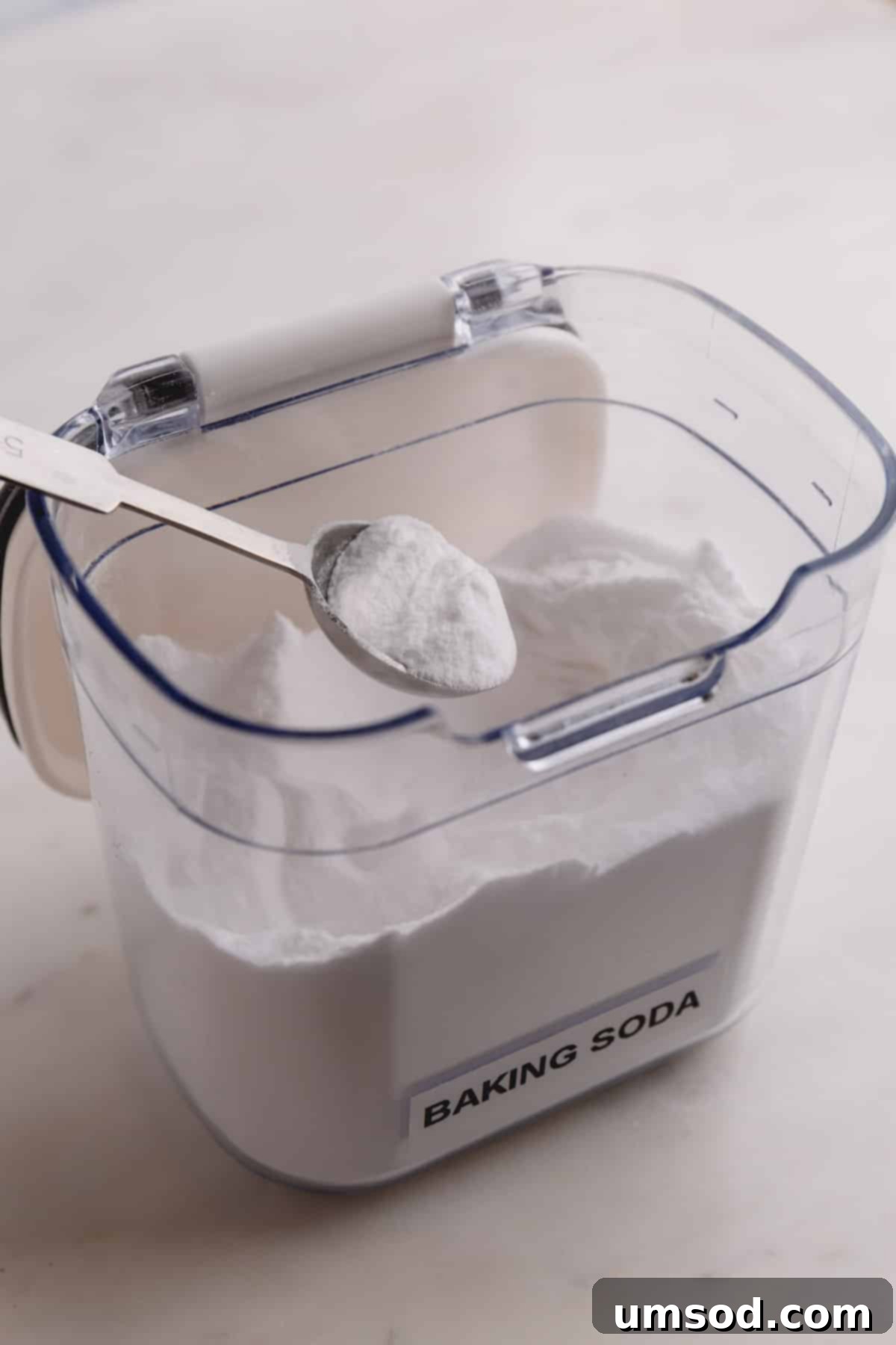Baking soda in an open container with a measuring spoon full of baking soda, highlighting its fine, white powder form.