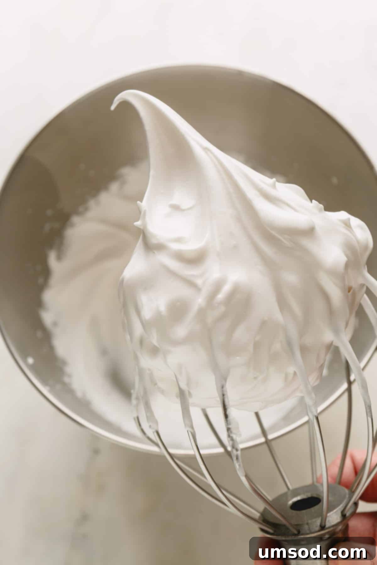 A whisk lifting fluffy vegan meringue from a mixing bowl, showcasing its perfect stiff peaks.