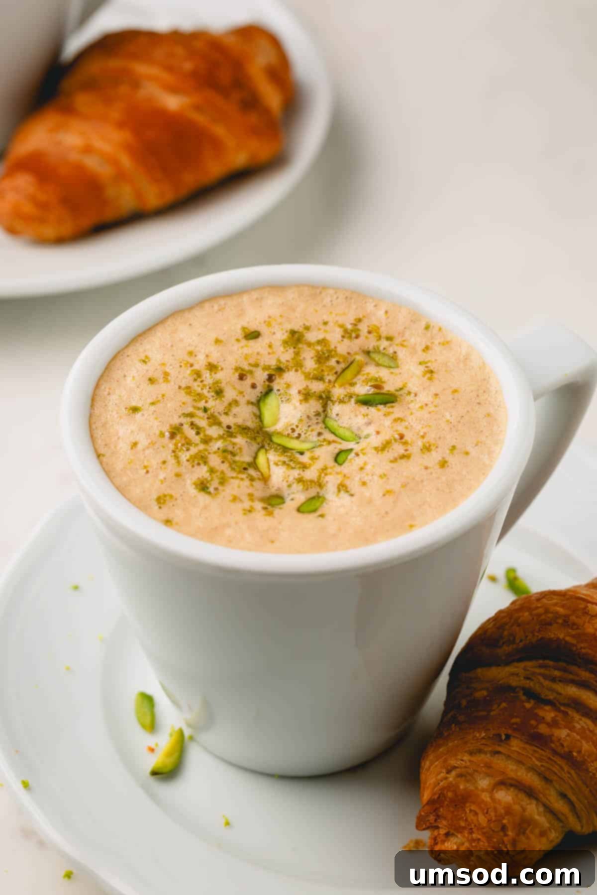 A beautifully crafted pistachio latte, garnished with crushed pistachios, served alongside a flaky croissant on a rustic plate.
