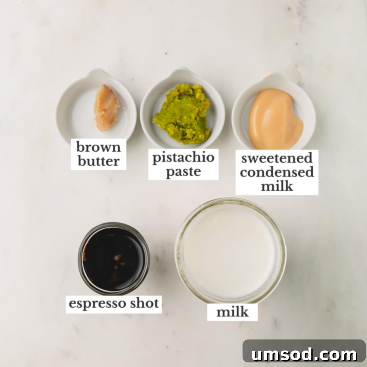 The five key ingredients for a homemade pistachio latte: brown butter, pistachio paste, sweetened condensed milk, espresso, and milk, artfully arranged on a white surface.