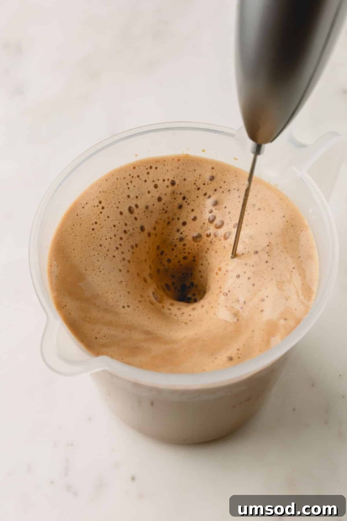 A handheld milk frother actively frothing a pistachio latte in a liquid measuring cup, creating a creamy texture.