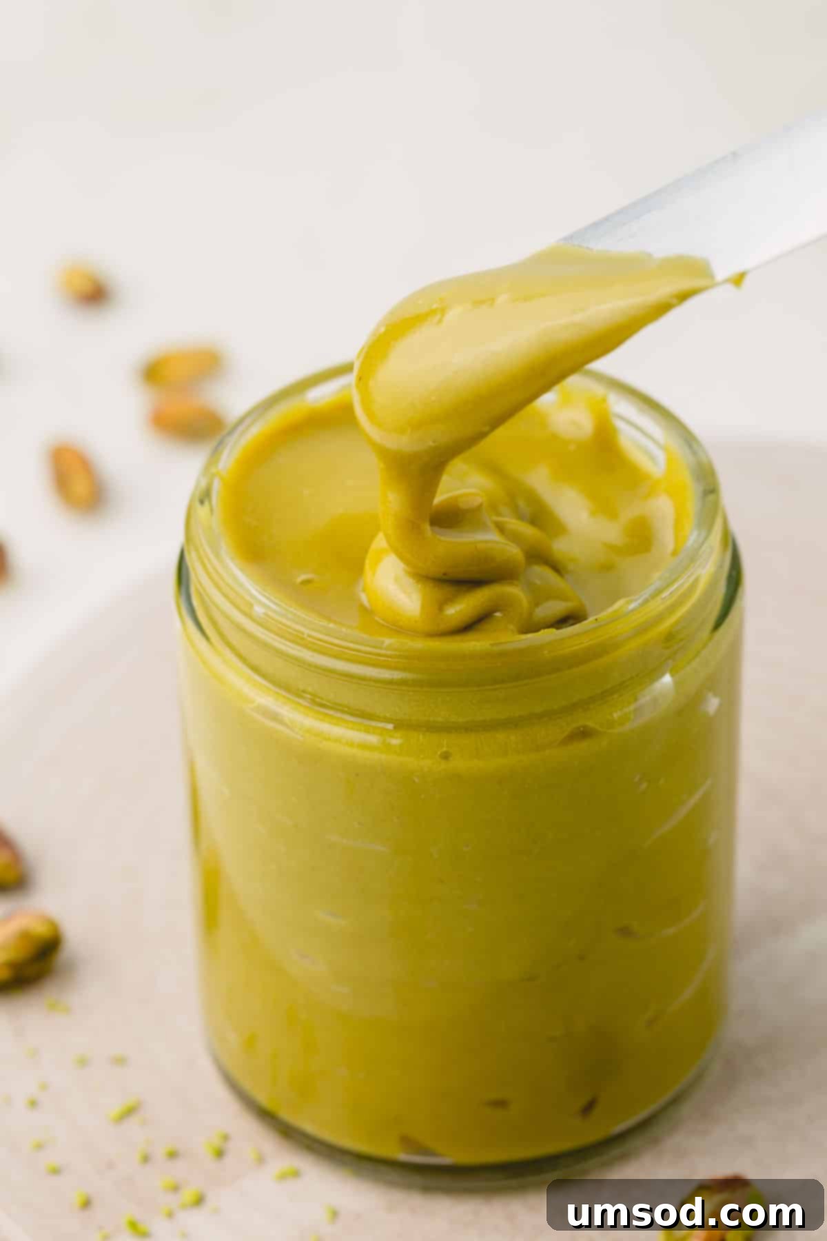A knife lifting a generous scoop of vibrant green pistachio butter from a clear glass jar.