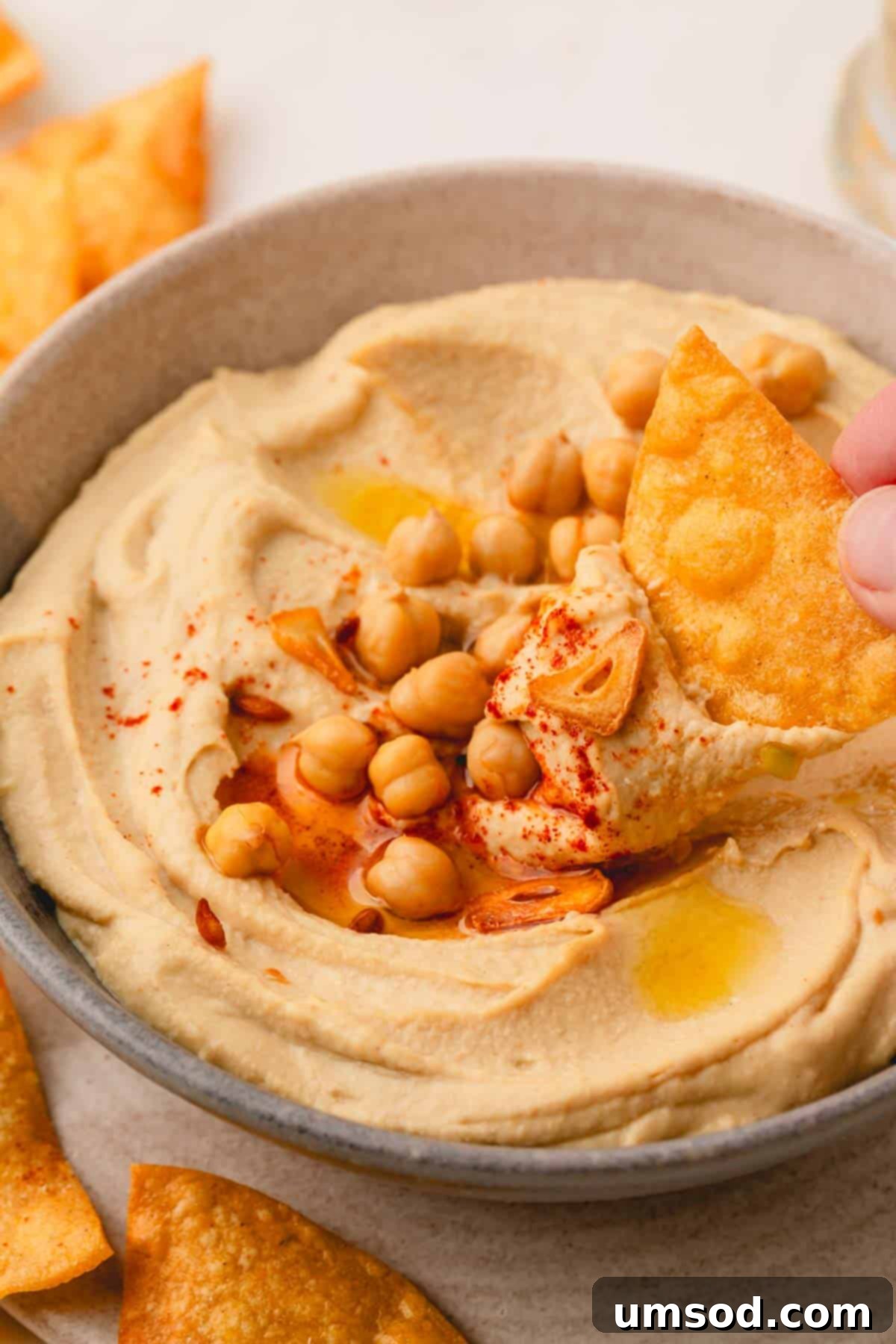 A crisp pita chip being dipped into a beautifully garnished bowl of roasted garlic hummus, highlighting its creamy texture.