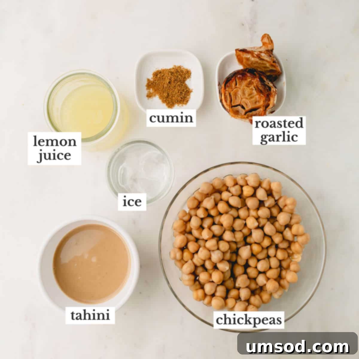 Bowls containing the key ingredients for roasted garlic hummus: lemon juice, cumin, roasted garlic cloves, ice cubes, tahini, and rinsed chickpeas.