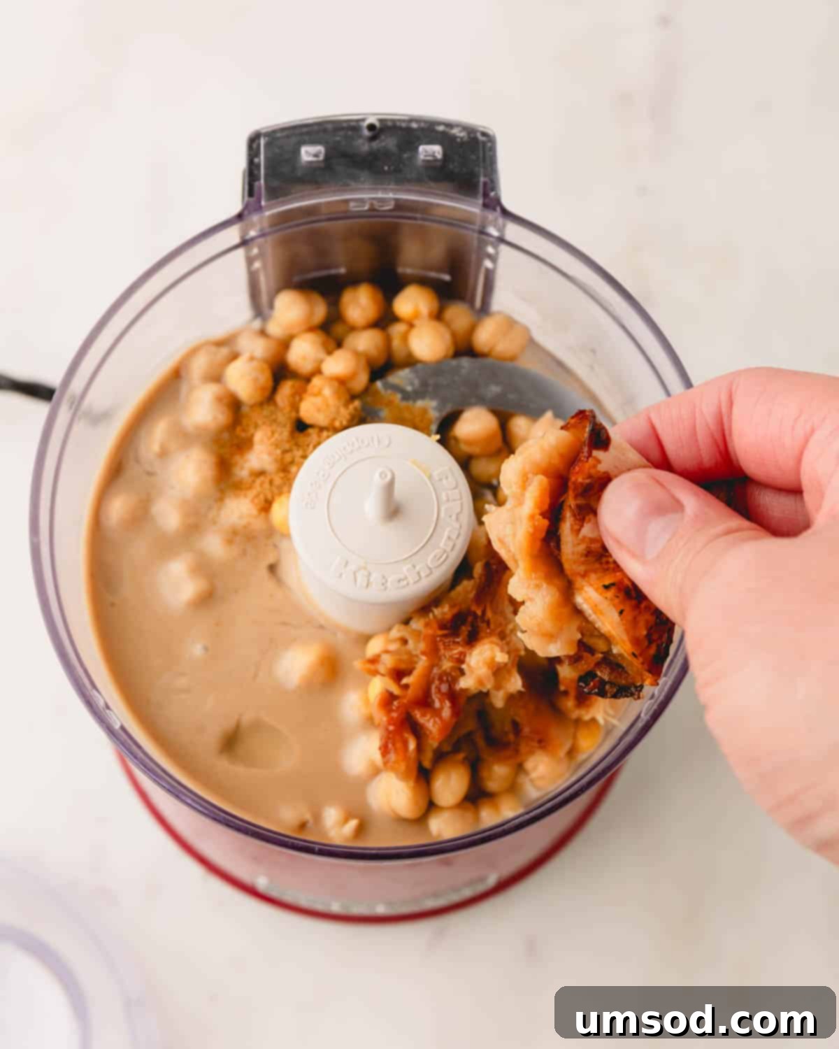 Soft roasted garlic cloves being squeezed directly into a food processor with other hummus ingredients like chickpeas and tahini.