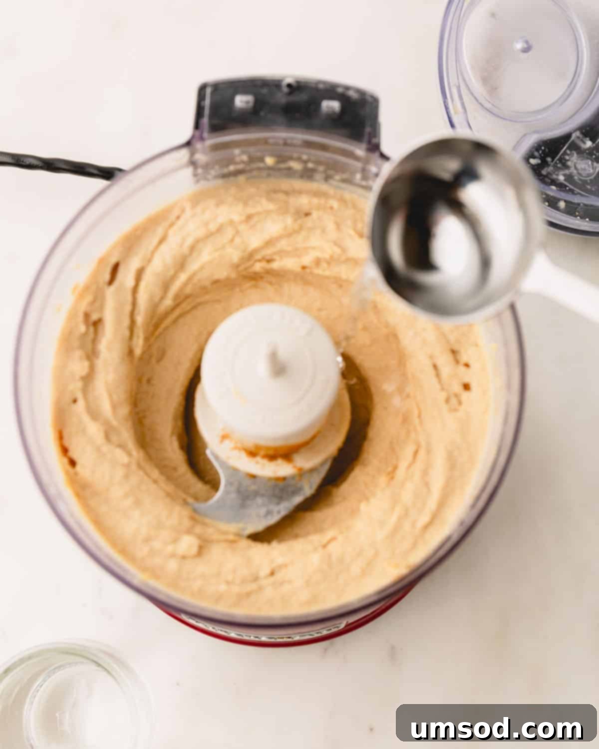 A thin stream of ice-cold water being carefully drizzled into a food processor while it blends the hummus ingredients.
