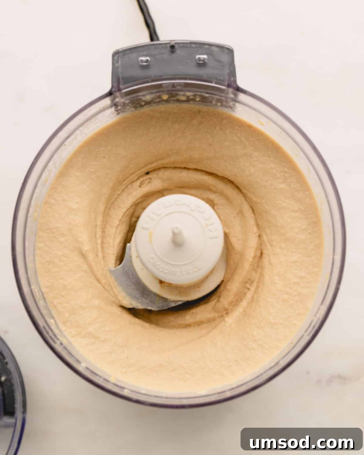 A close-up of a bowl of beautifully smooth roasted garlic hummus inside a food processor, ready to be served.