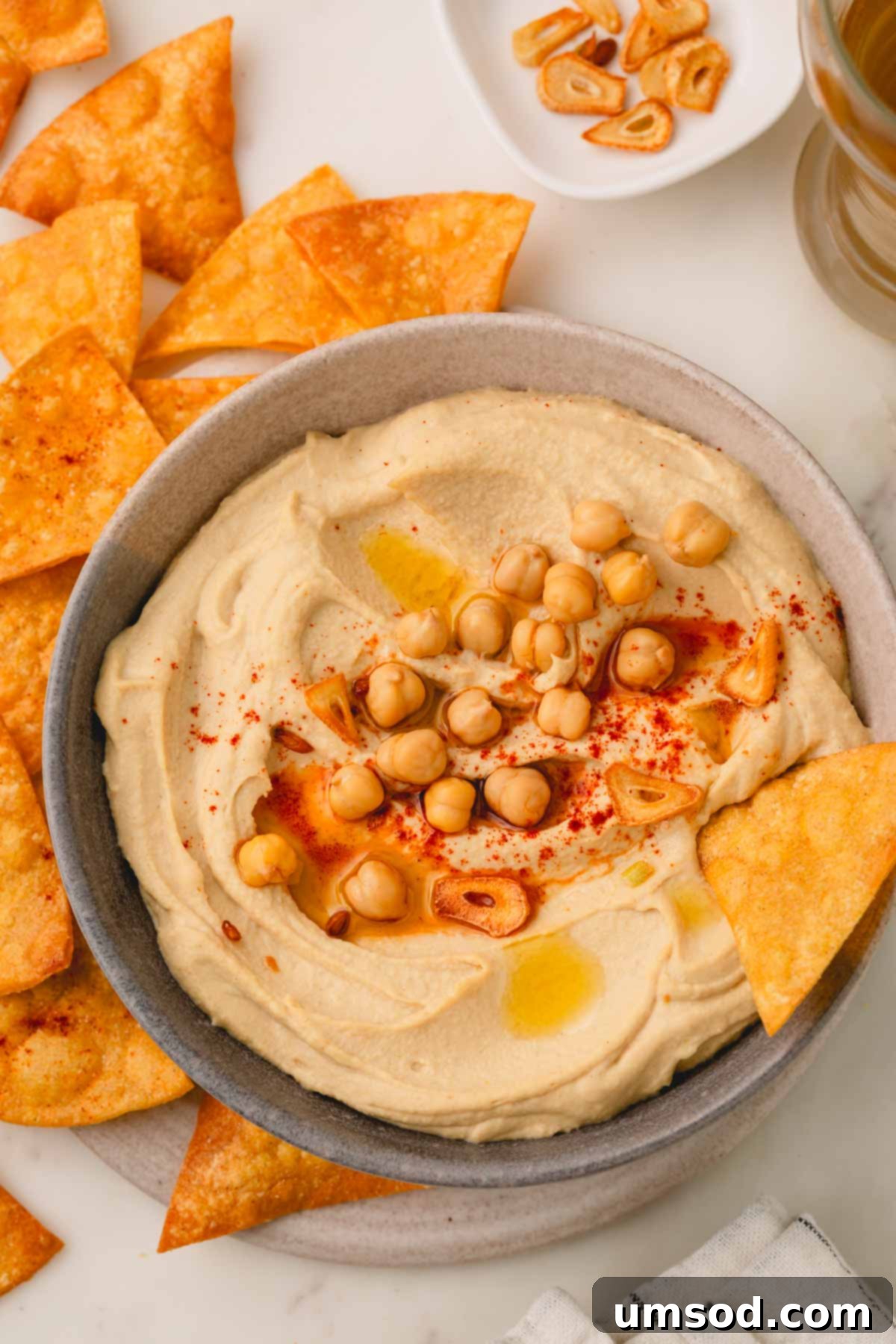 A generous bowl of roasted garlic hummus, garnished with a swirl of olive oil and paprika, with a pita chip ready for dipping and a pile of chips beside it.