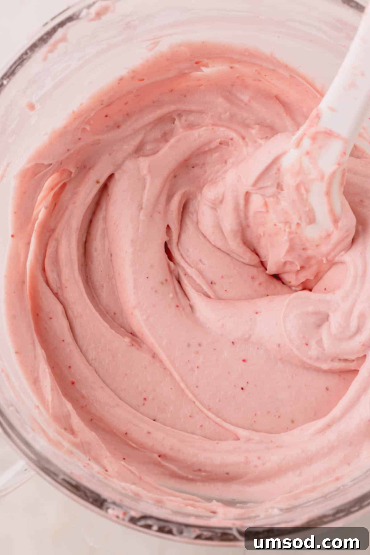 A large bowl of smooth, pink strawberry cream cheese frosting with a spatula resting inside it, ready for use.