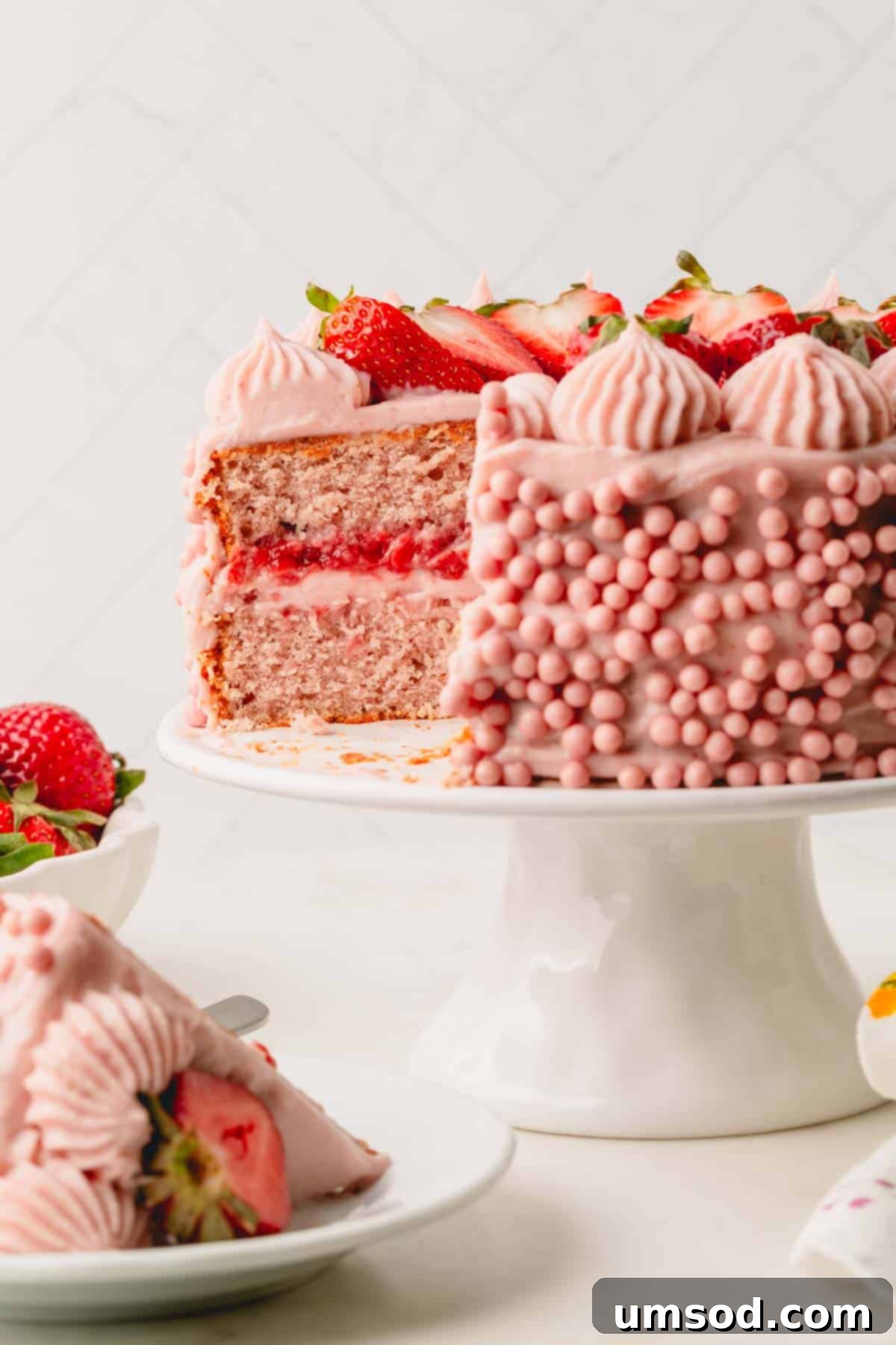 Berry Sweet Symphony 2 A two layer frosted strawberry cake on a cake stand with a slice missing.