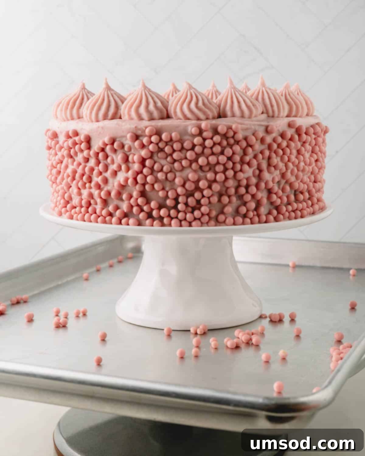Berry Sweet Symphony 18 A strawberry cake on a cake stand on a baking sheet with strawberry pearls on the side.