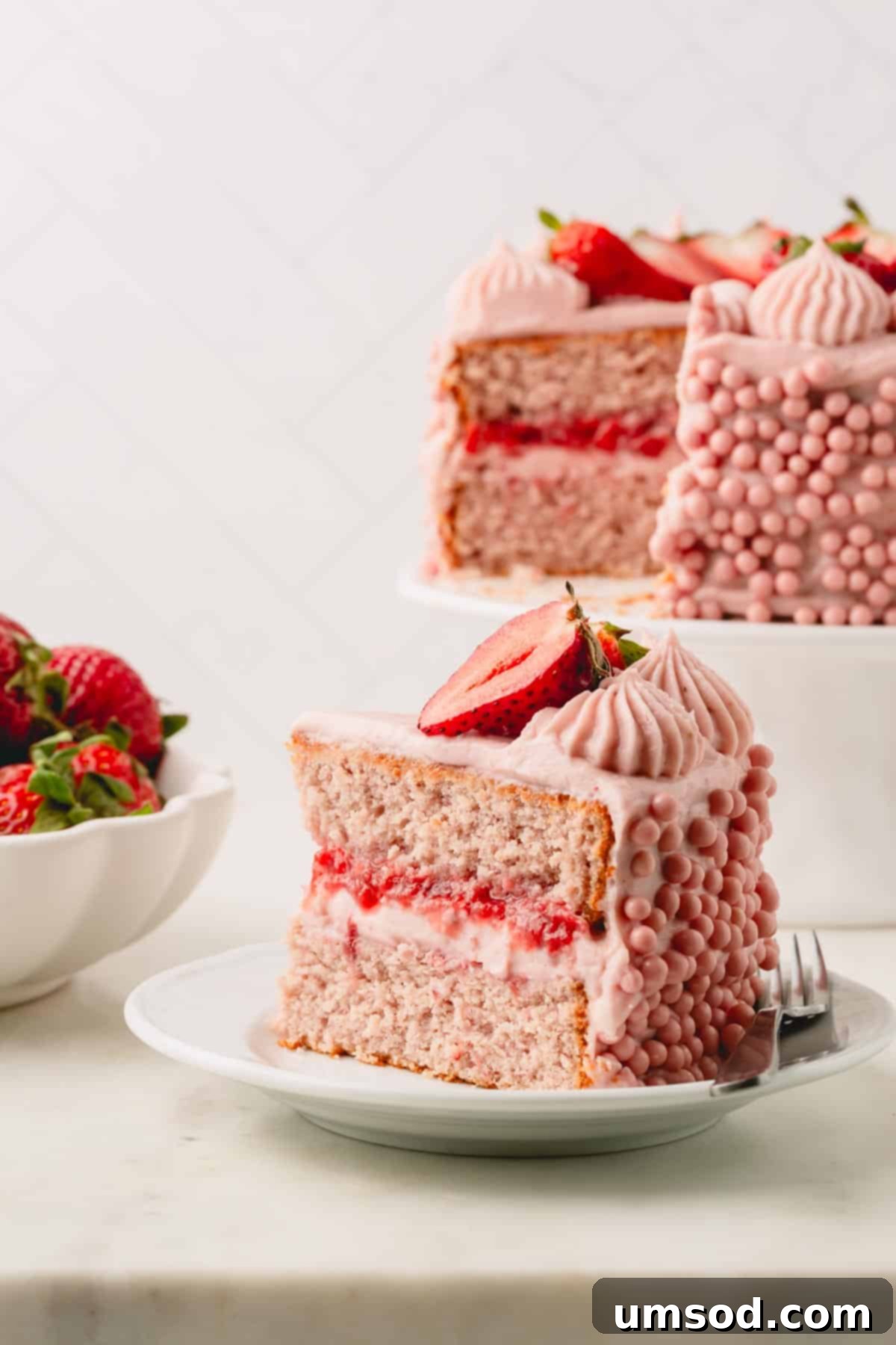 Berry Sweet Symphony 3 A slice of layered frosted strawberry cake on a plate next to the rest of the cake on the stand.