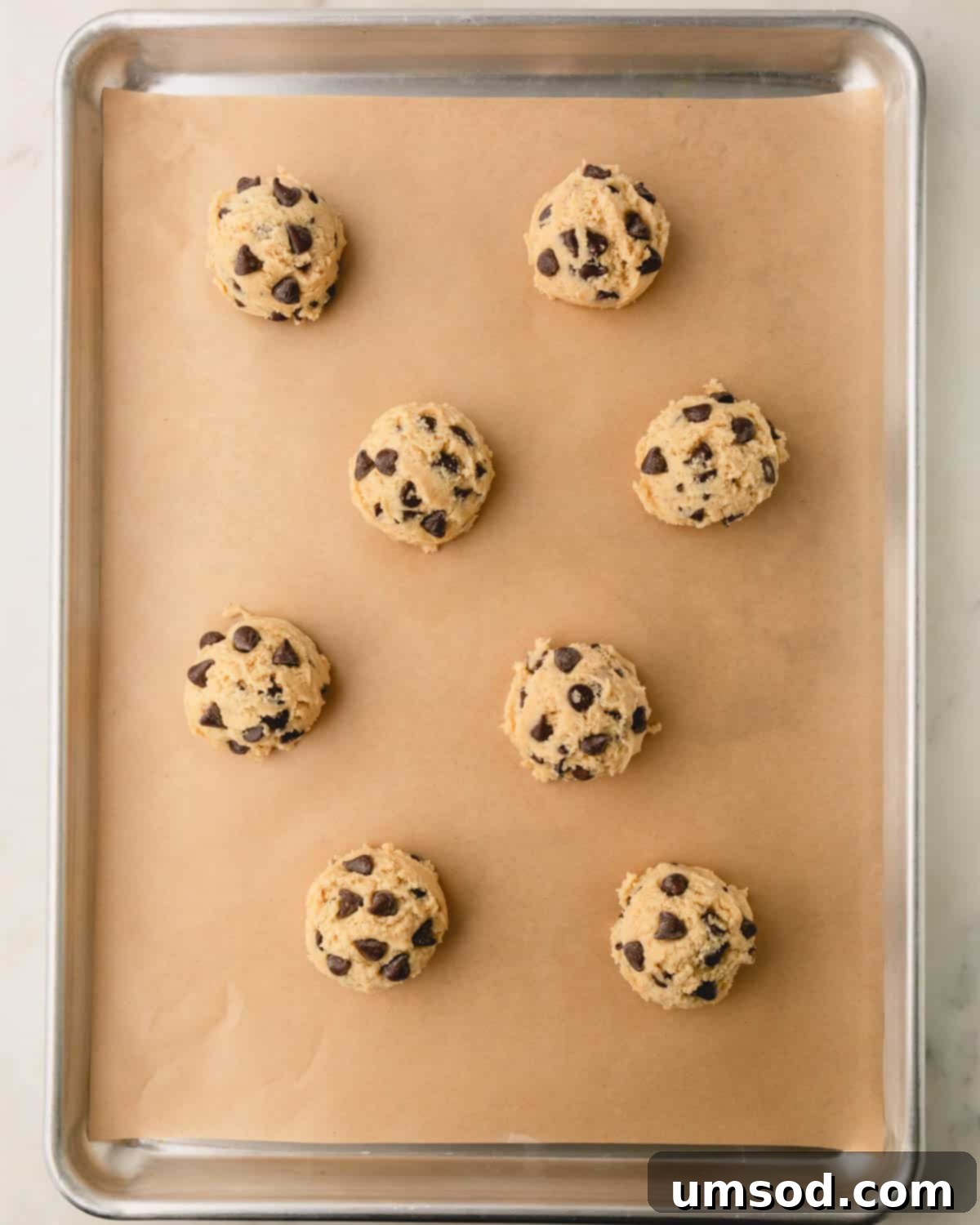 Raw chocolate chip cookie dough balls on a parchment paper-lined baking sheet.