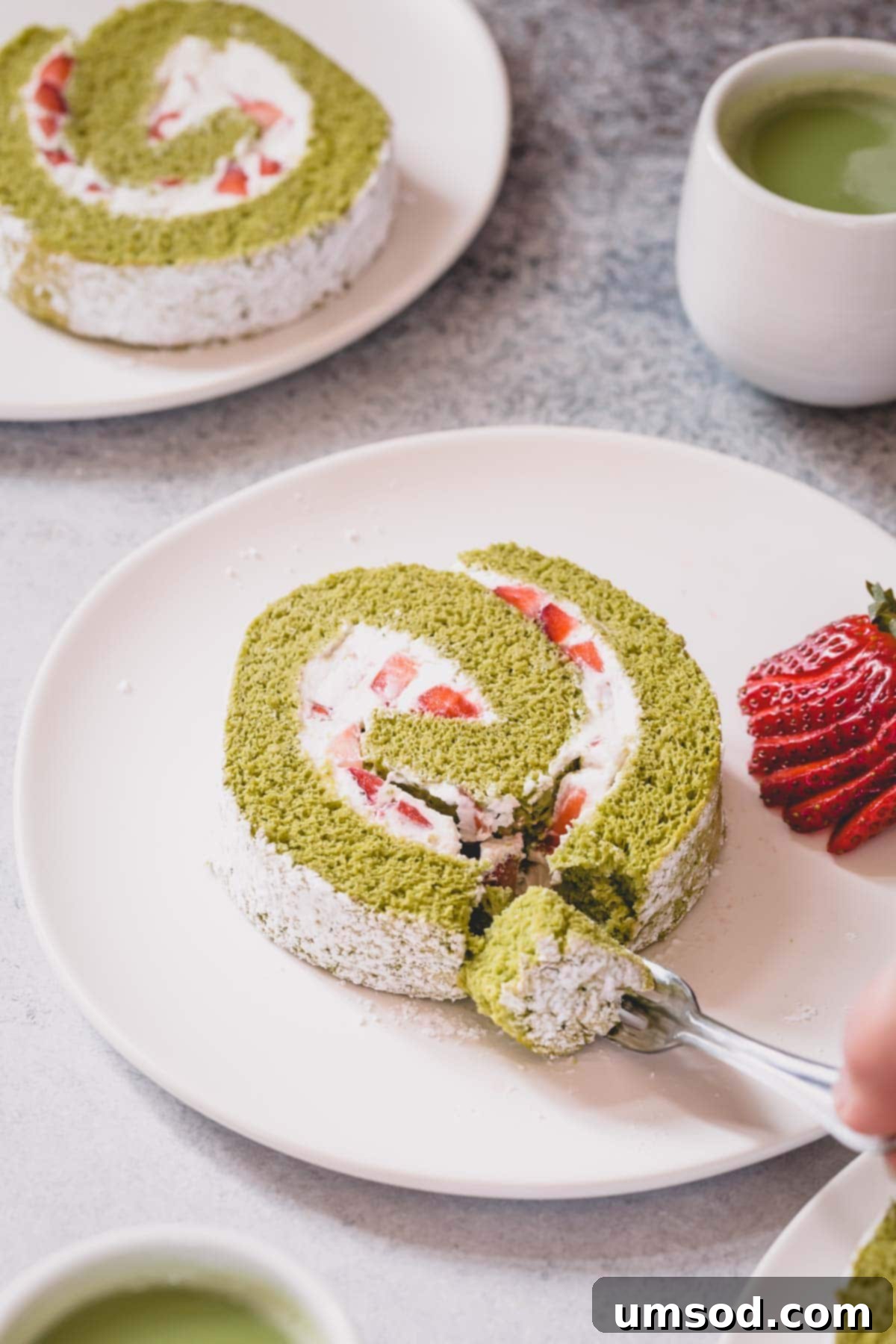 Matcha Strawberry Swirl Cake 3 A slice of strawberry matcha roll cake on a white serving plate with a a little slice cut with a fork.